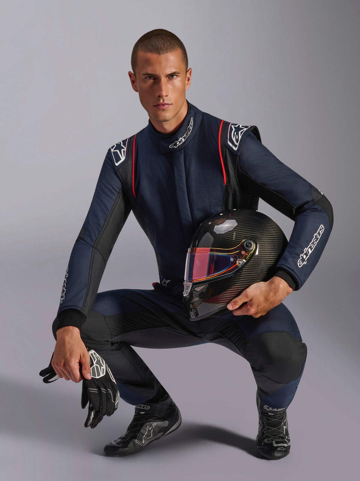 Pilota automobilistico con tuta Alpinestars GP Tech V4, blu navy con accenti neri e rossi, regge un casco in fibra di carbonio con visiera iridescente, guanti e stivaletti da gara neri, abbigliamento professionale per competizioni automobilistiche