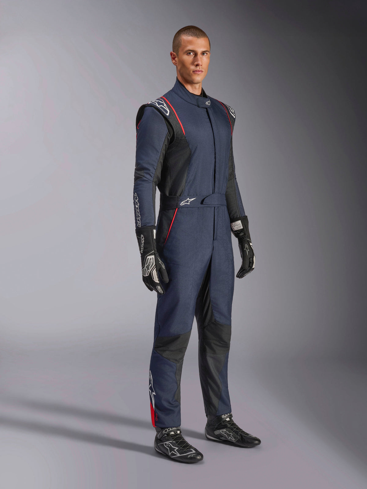 Tuta Alpinestars GP Tech V4, tuta intera da auto racing, blu navy, nero e rosso, indossata da un modello con guanti e stivali da corsa neri, caratterizzata da pannelli neri e profili rossi, abbigliamento professionale per motorsport.