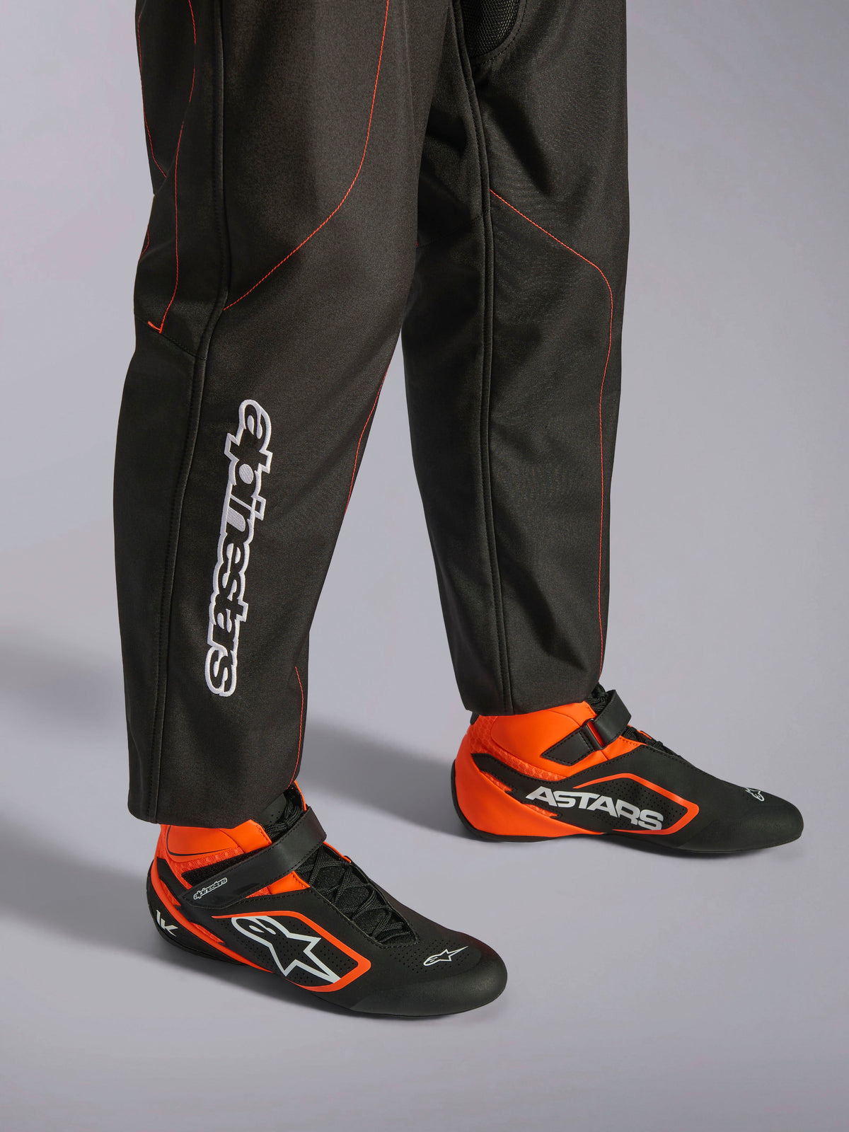 Tuta Alpinestars KMX-9 V3, tuta da kart, nero e rosso fluo, vista dettagliata della parte inferiore delle gambe e delle scarpe da corsa, tessuto nero con cuciture rosso fluo, logo Alpinestars verticale sul polpaccio, abbinata a stivaletti da corsa neri e arancioni coordinati, progettata per il karting professionale