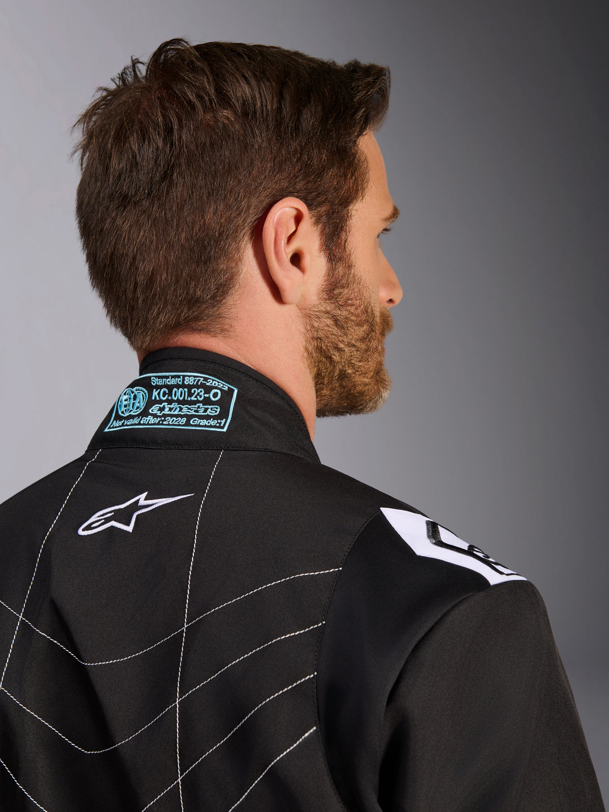 Tuta Alpinestars KMX-9 V3, nero e bianco, primo piano del colletto posteriore e della zona delle spalle, con dettagli dell'omologazione FIA ricamati in azzurro, logo a stella bianco e cuciture bianche a contrasto