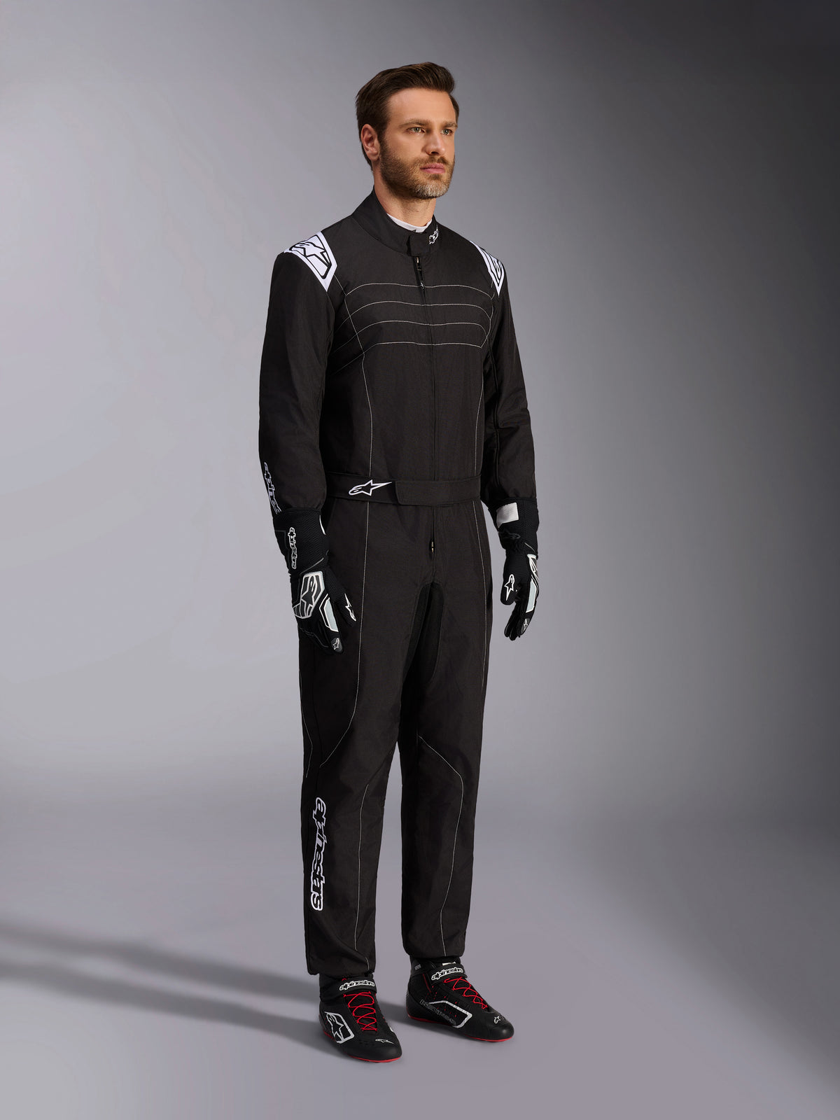 Tuta da karting Alpinestars KMX-9 V3, nero e bianco, modello maschile che indossa la tuta da corsa completa con logo Alpinestars bianco sulle spalline, abbinata a guanti da corsa bianchi e neri e scarpe da kart nere con lacci rossi, scatto in studio che mostra il profilo frontale e laterale