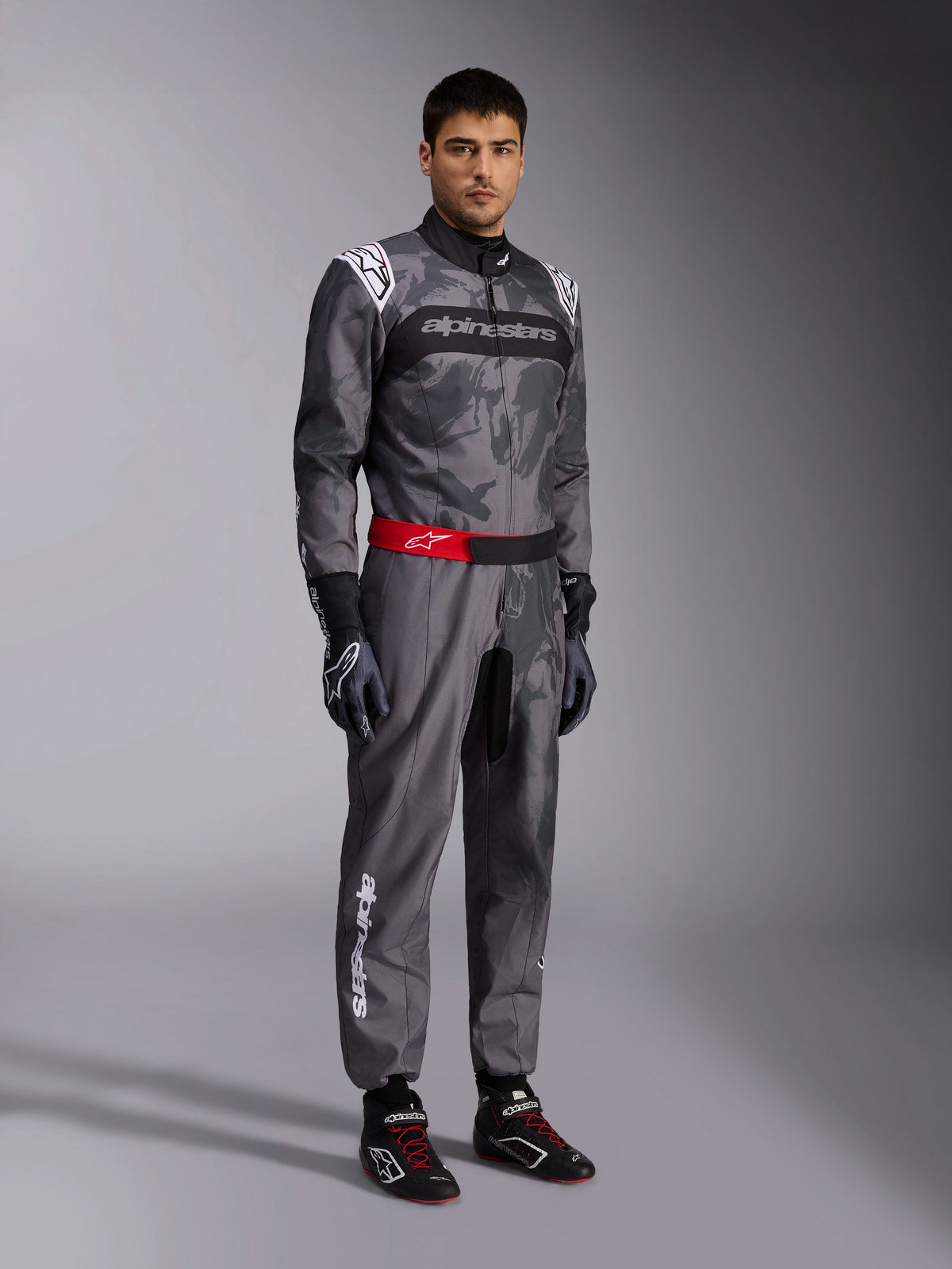 Alpinestars Tuta KMX-9 V3 Graphic 5, tuta da karting, grigio scuro e nero con motivo grafico mimetico, cintura rossa in vita con logo bianco, indossata con guanti e scarpe da corsa, abbigliamento professionale per karting
