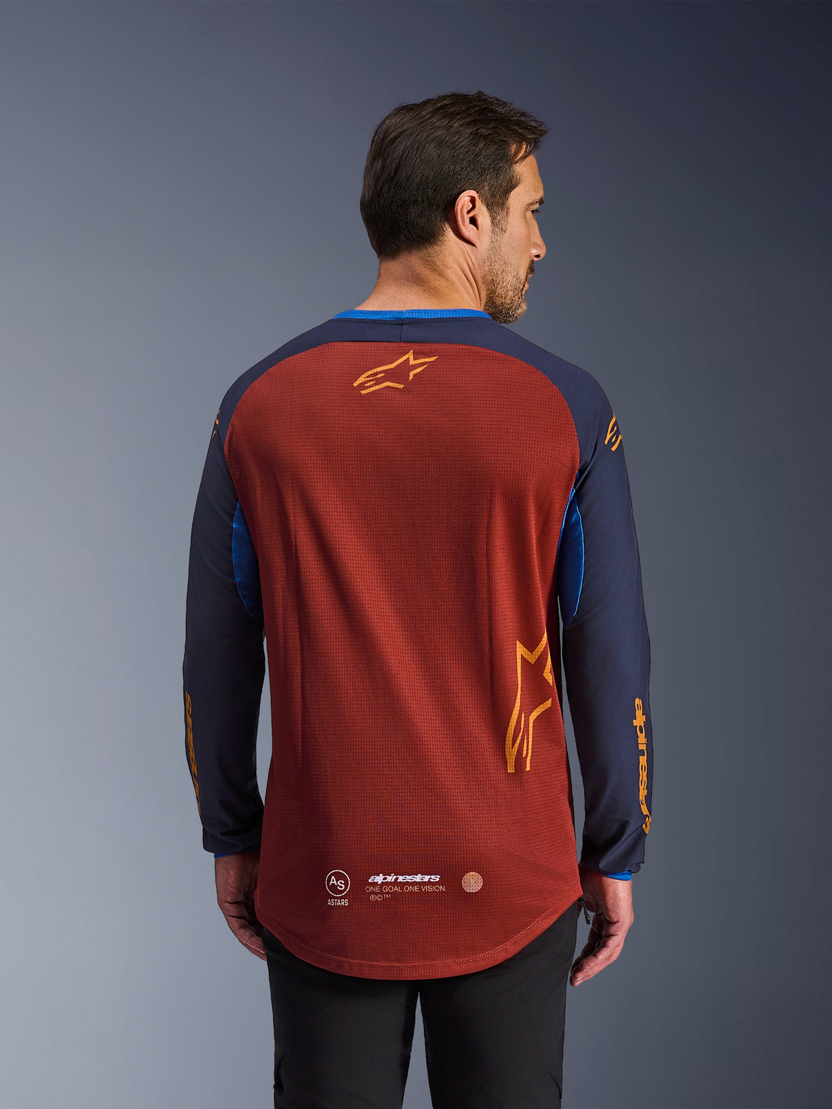 Alpinestars Maxdura Dual Jersey in Rust Brown e Mood Indigo, vista dal retro, caratterizzata da busto in mesh traspirante, maniche blu scuro con logo Alpinestars arancione e orlo posteriore allungato per una maggiore copertura.