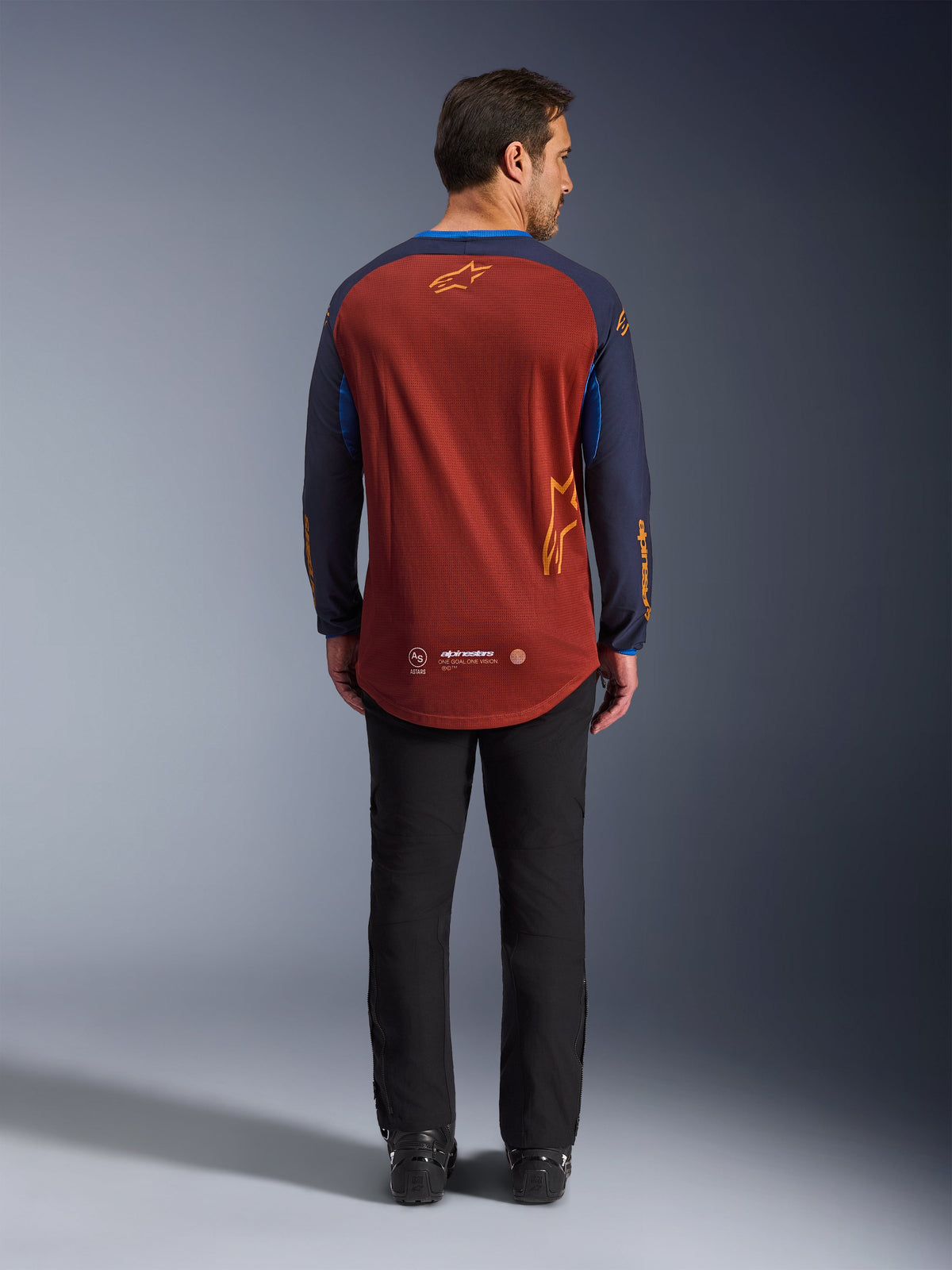 Alpinestars Maxdura Dual Jersey in Rust Brown e Mood Indigo, vista posteriore che mostra l'orlo posteriore allungato e i dettagli del logo, indossata con pantaloni tecnici neri e stivali, progettata per il motociclismo d'avventura e dual-sport.