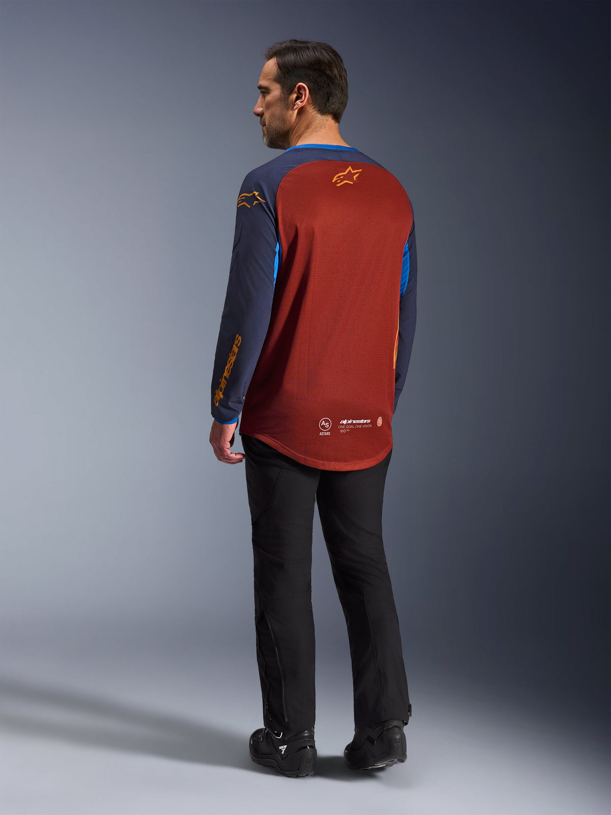 Alpinestars Maxdura Dual Jersey, Rust Brown e Mood Indigo, maglia da moto adventure vista dal retro, pannello in mesh color ruggine per la ventilazione, maniche blu navy con loghi arancioni, indossata con pantaloni e stivali da moto neri, abbigliamento tecnico da fuoristrada