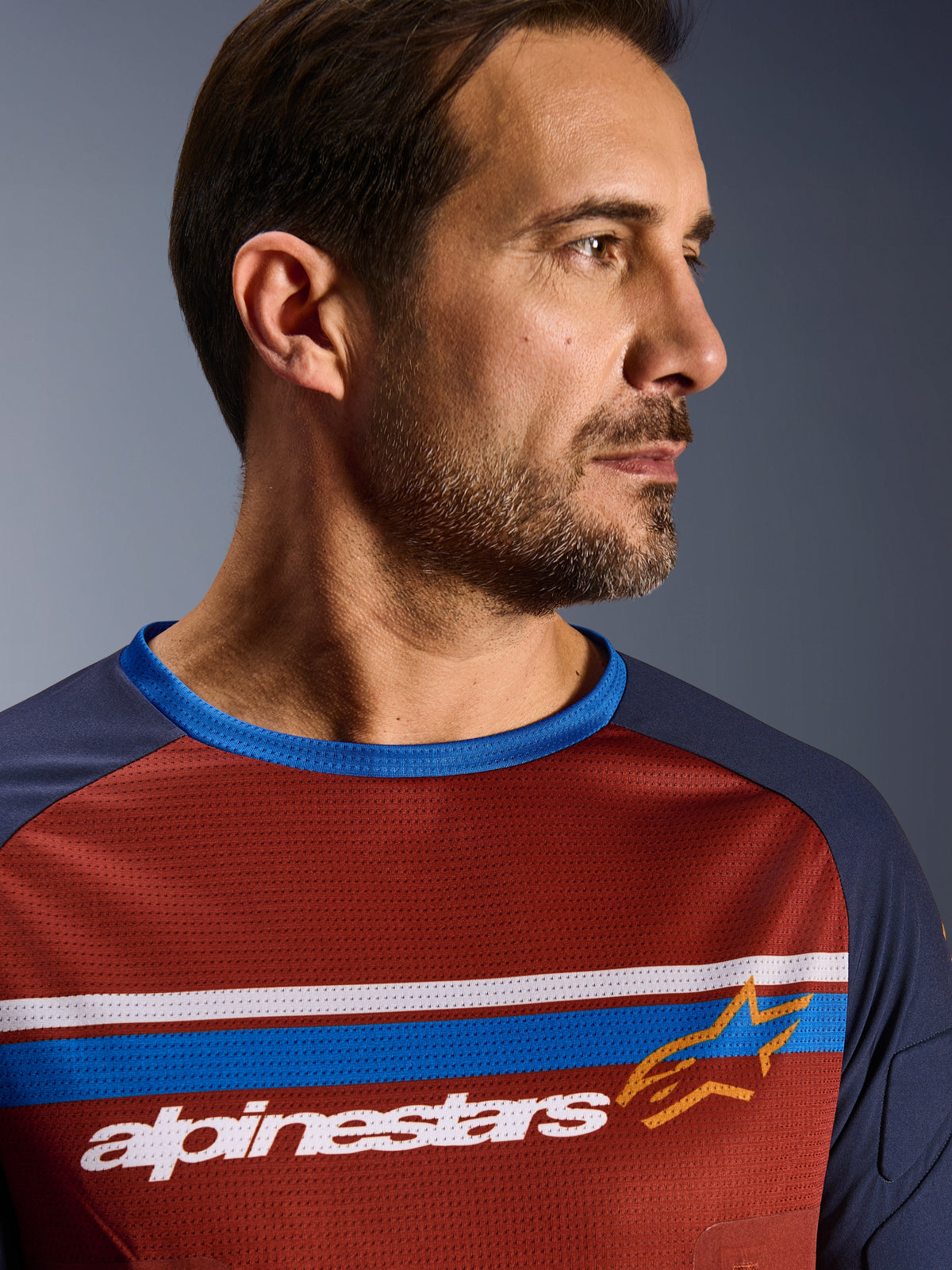 Alpinestars Maxdura Dual Jersey, Rust Brown e Mood Indigo, primo piano di un pilota che indossa la maglia tecnica in mesh, caratterizzata da strisce orizzontali bianche e blu sul petto, marchio Alpinestars bianco e logo a stella arancione.