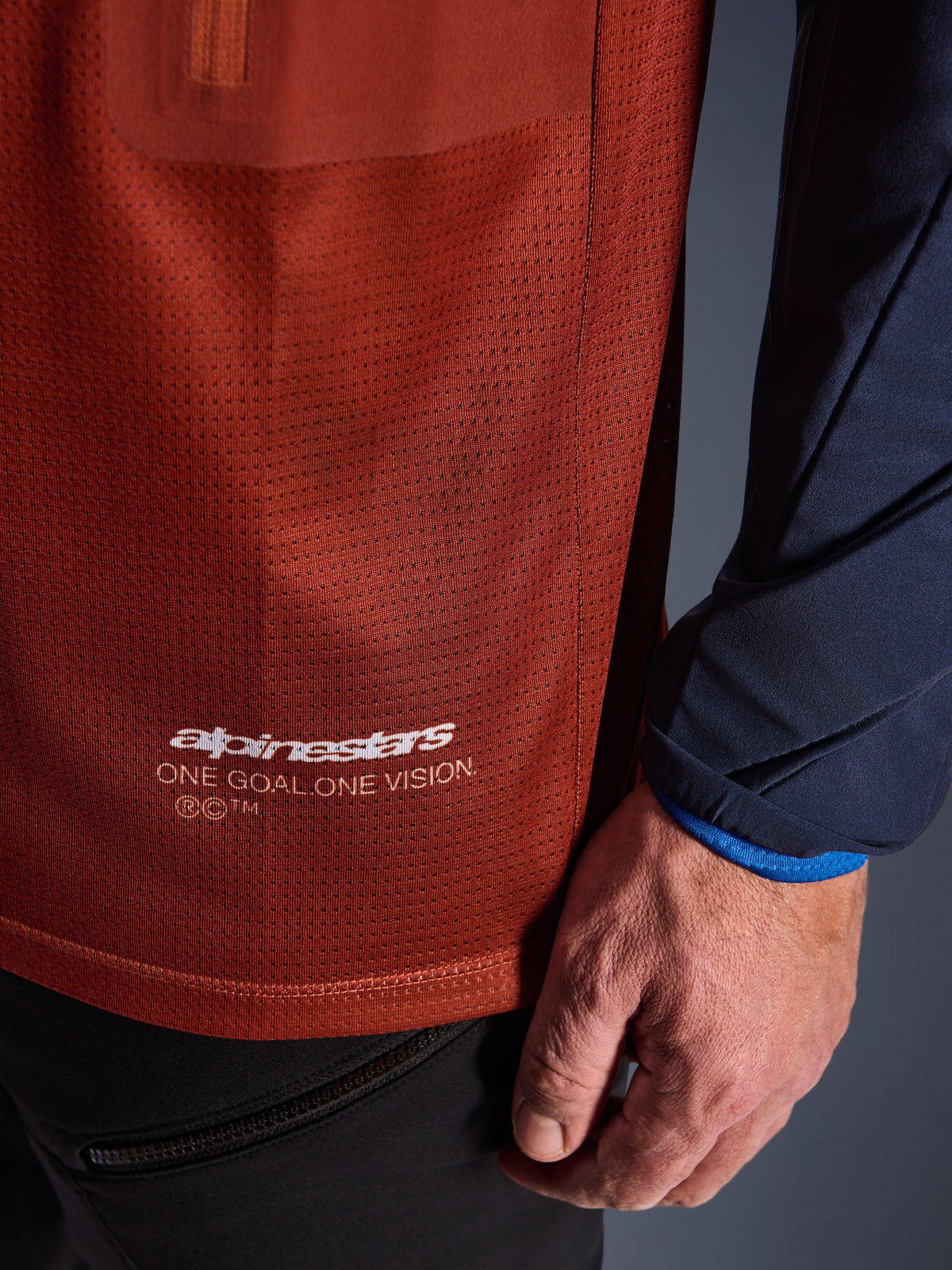 Alpinestars Maxdura Dual Jersey, maglia da moto adventure, marrone ruggine e blu Mood Indigo, vista ravvicinata che mostra il tessuto a rete del busto e la manica in tessuto blu scuro, logo Alpinestars 'One Goal One Vision' bianco sull'orlo inferiore