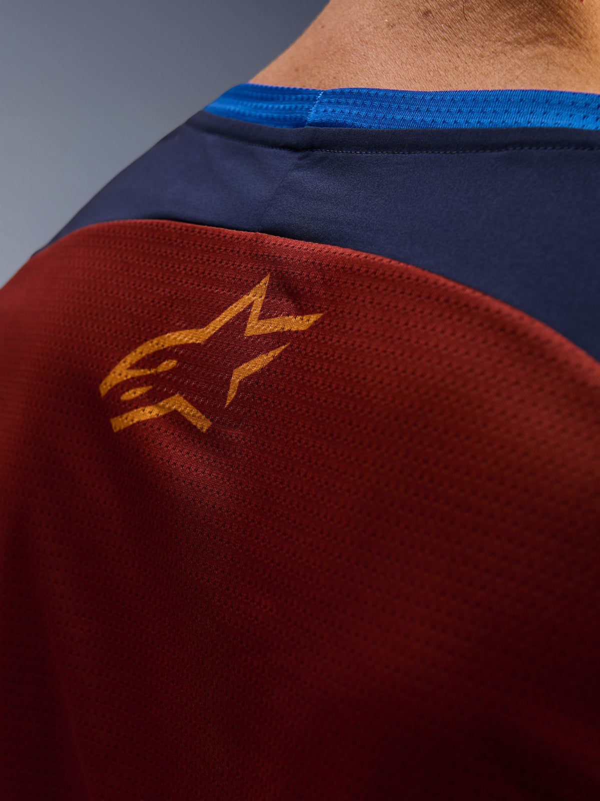 Primo piano della maglia Alpinestars Maxdura Dual Jersey, tessuto a rete marrone ruggine sul retro, piccolo logo a stella arancione, pannello superiore blu scuro e bordo del colletto blu acceso, vista dettagliata maglia moto adventure