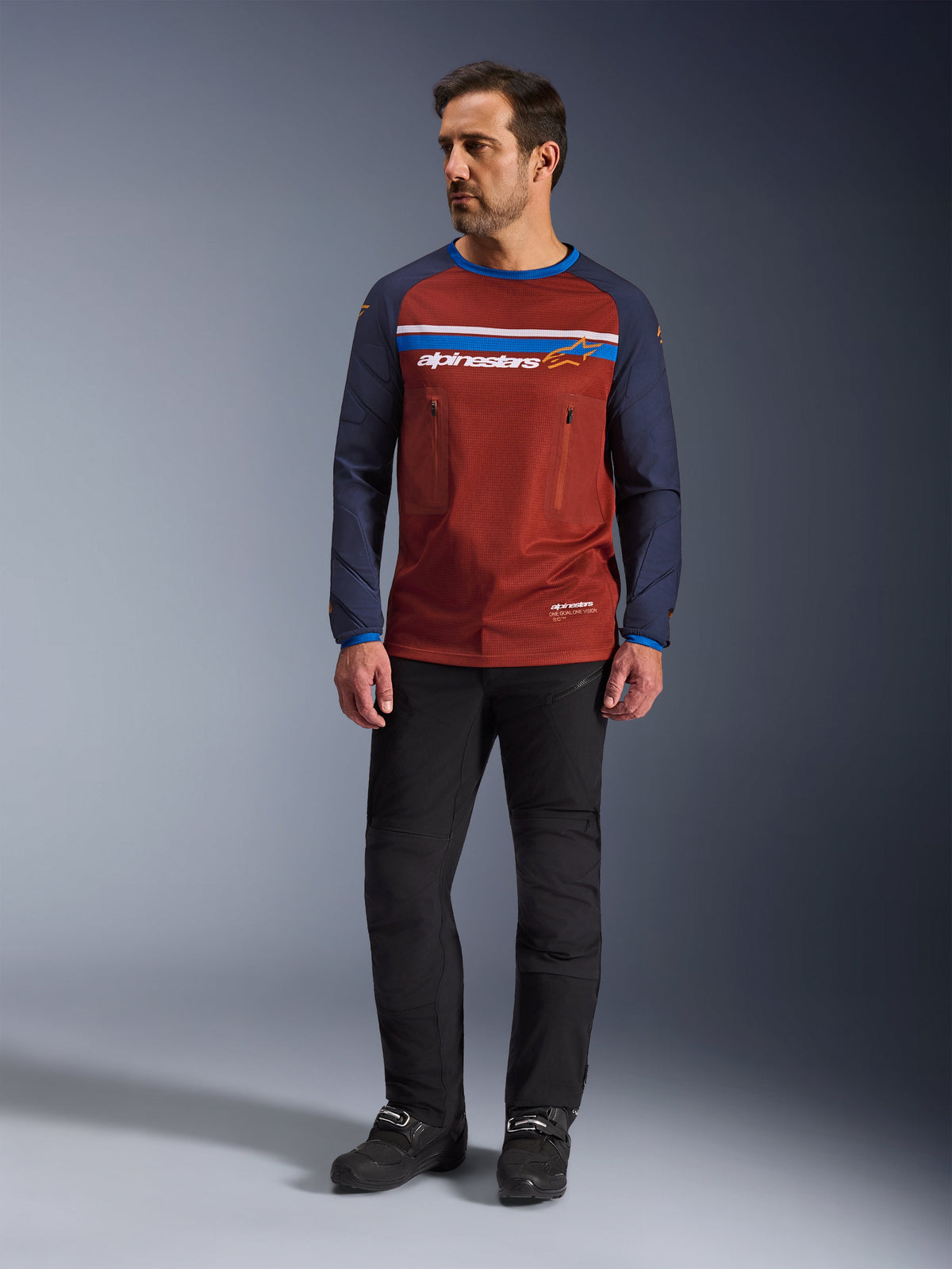 Alpinestars Maxdura Dual Jersey, Rust Brown e Mood Indigo, maglia da moto adventure con busto in rete e maniche blu navy, indossata da un modello con pantaloni da guida neri e stivali neri, abbigliamento tecnico dual-sport per la guida off-road