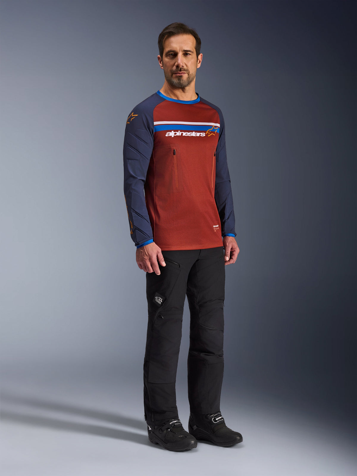 Uomo che indossa la maglia Alpinestars Maxdura Dual Jersey in Rust Brown e Mood Indigo, con busto in rete e maniche imbottite, abbinata a pantaloni da moto e stivali neri, abbigliamento da moto adventure.
