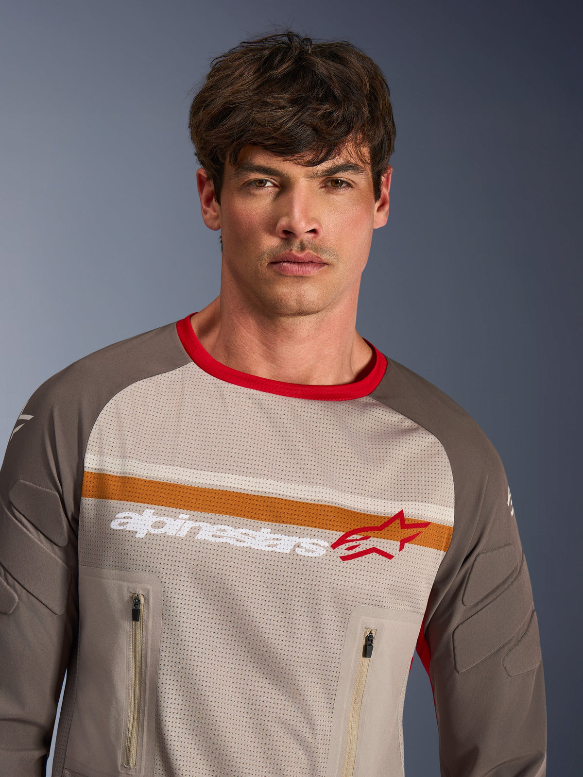 Modello che indossa Alpinestars Maxdura Dual Jersey in Aluminum Walnut, maglia da moto adventure con torso completamente in rete, maniche resistenti all'abrasione, colletto rosso e tasche sul petto con cerniera, con strisce arancioni e bianche sul petto e logo Alpinestars.