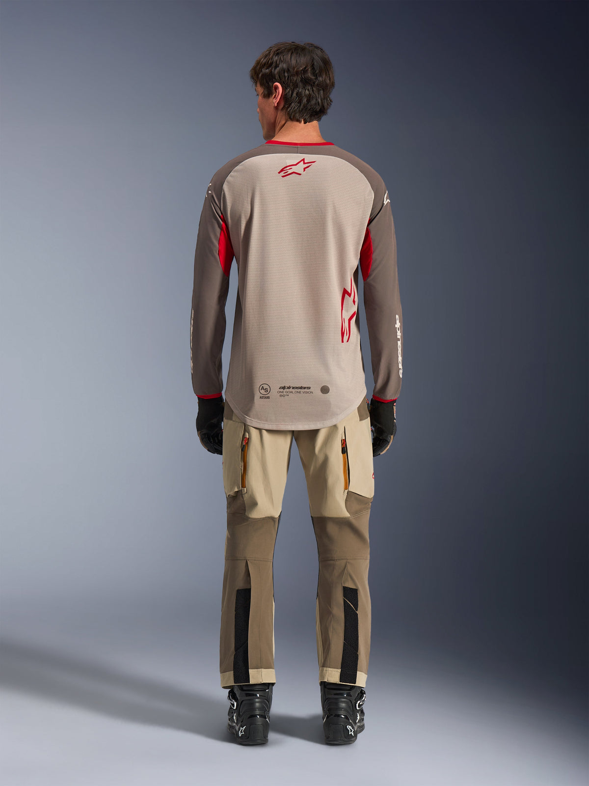 Alpinestars Maxdura Dual Jersey in Aluminum Walnut, vista posteriore, torso in mesh color sabbia con maniche marroni e loghi rossi, bordo posteriore allungato, abbinata a pantaloni da adventure e stivali neri.