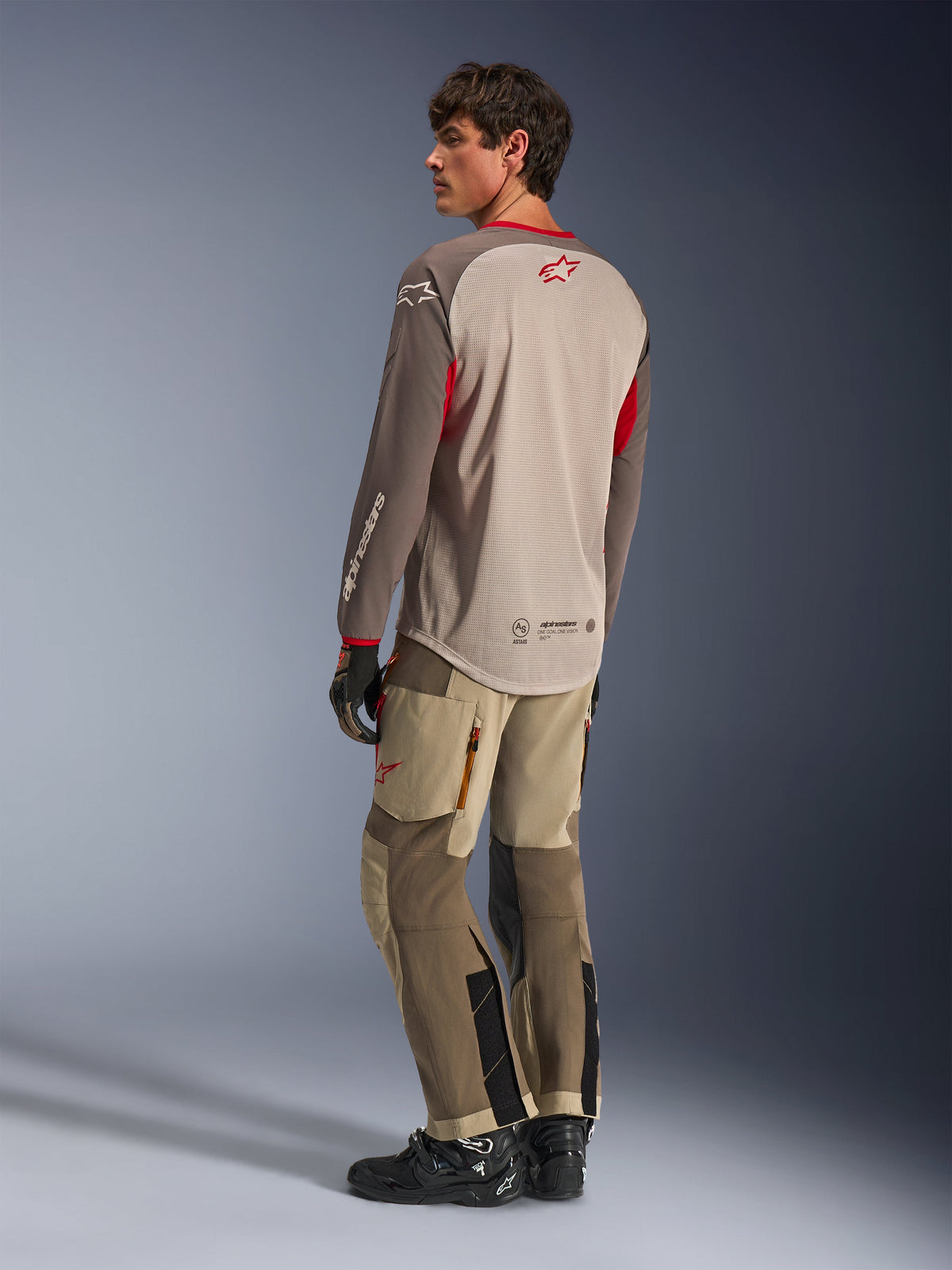 Alpinestars Maxdura Dual Jersey in Aluminum Walnut, vista posteriore della maglia da moto adventure con busto in rete traspirante e dettagli rossi, abbinata a pantaloni tecnici adventure coordinati e stivali neri