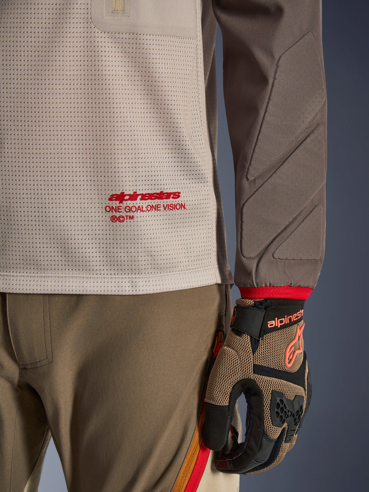 Alpinestars Maxdura Dual Jersey in Aluminum Walnut, vista dettagliata che mostra il busto in rete ventilata con logo rosso, manica resistente all'abrasione con imbottitura integrata, indossata con pantaloni e guanti da moto adventure coordinati