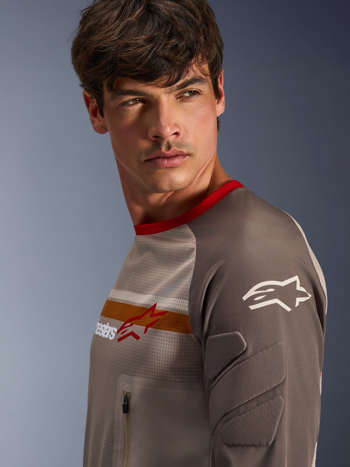 Modello maschile che indossa Alpinestars Maxdura Dual Jersey in Aluminum Walnut, con busto in mesh ventilato, colletto rosso e striscia arancione sul petto. Vista ravvicinata della manica resistente all'abrasione con imbottitura integrata e tasca sul petto con zip per guida adventure.