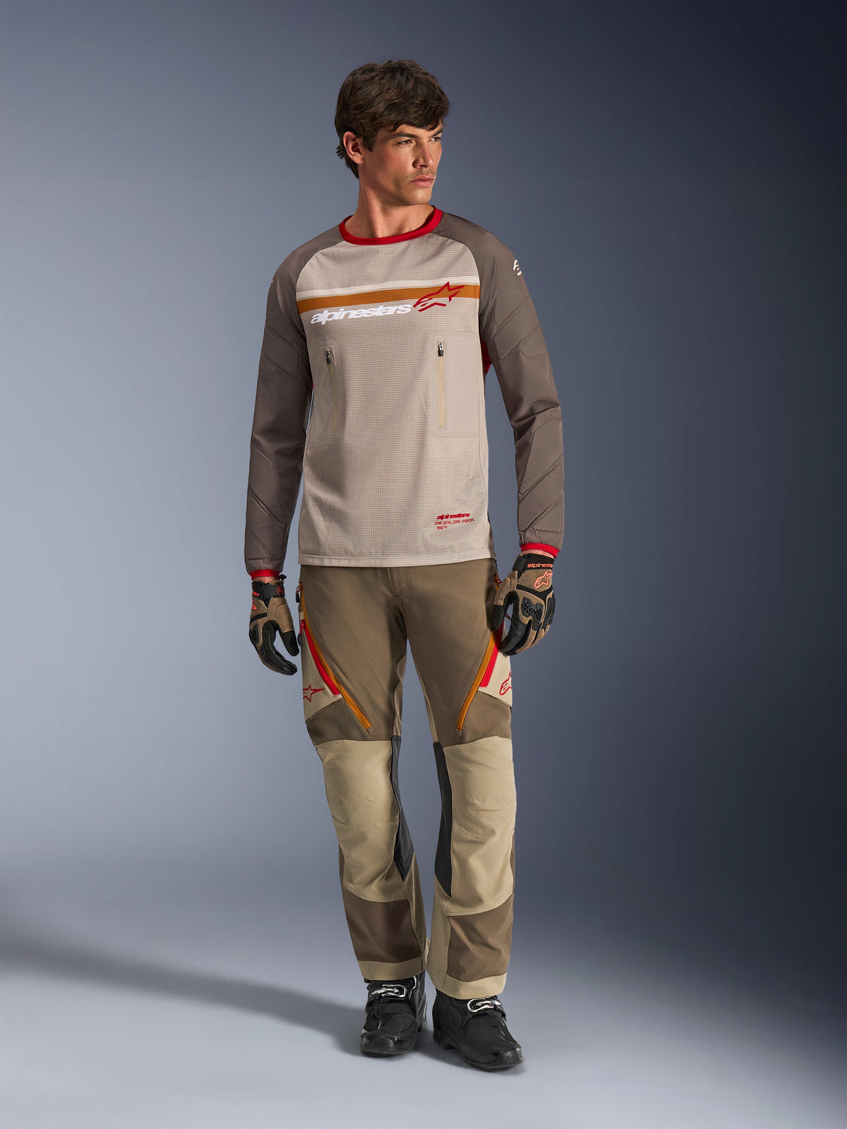 Alpinestars Maxdura Dual Jersey in Aluminum Walnut, maglia da moto adventure con busto in mesh color sabbia e maniche marroni, caratterizzata da colletto rosso e striscia orizzontale sul petto con logo Alpinestars, indossata con pantaloni e guanti adventure coordinati.