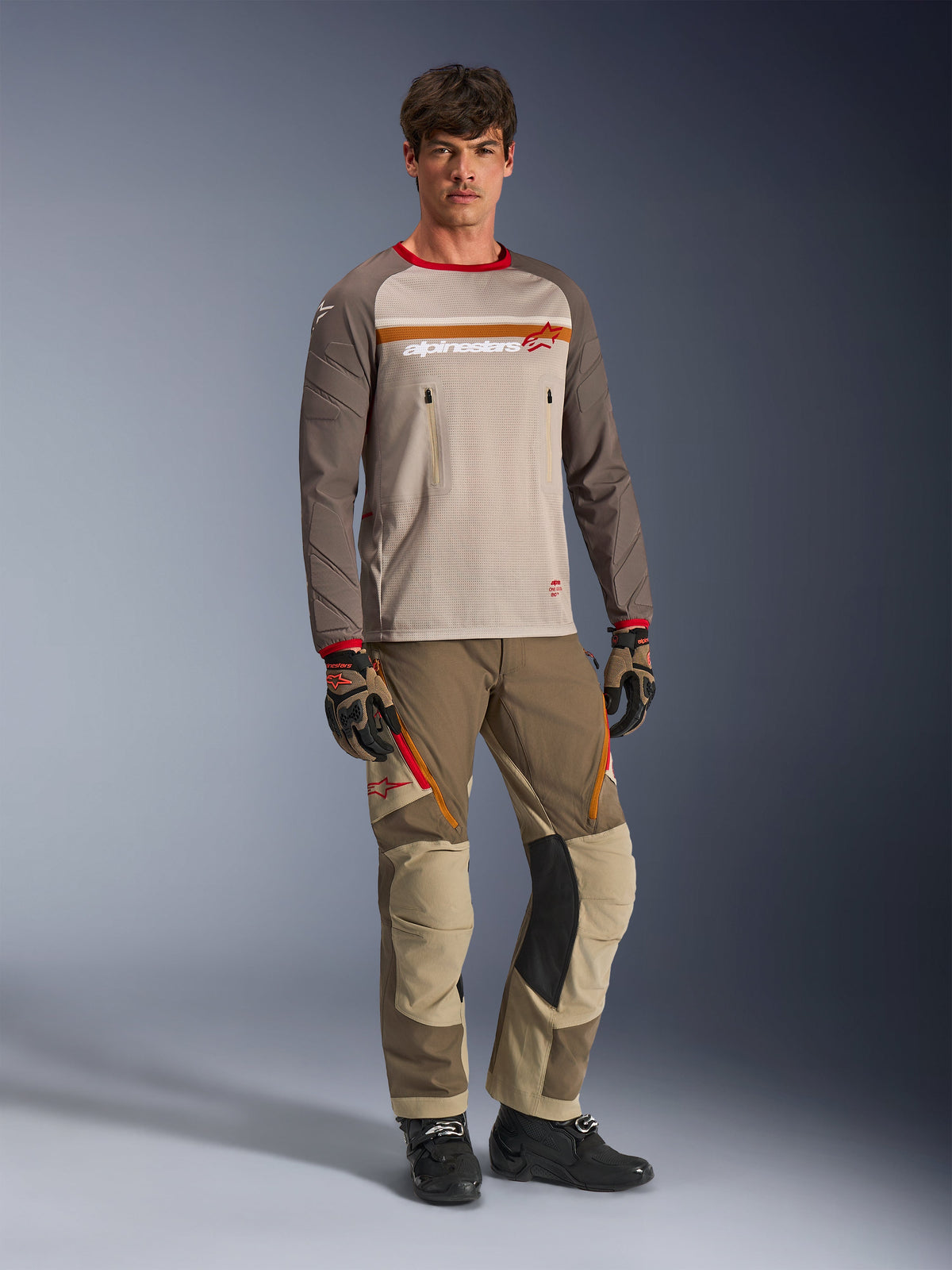 Alpinestars Maxdura Dual Jersey in Aluminum Walnut, maglia con busto in rete grigia, maniche marroni e polsini rossi, tasche sul petto con zip, indossata con abbigliamento tecnico coordinato per guida adventure.