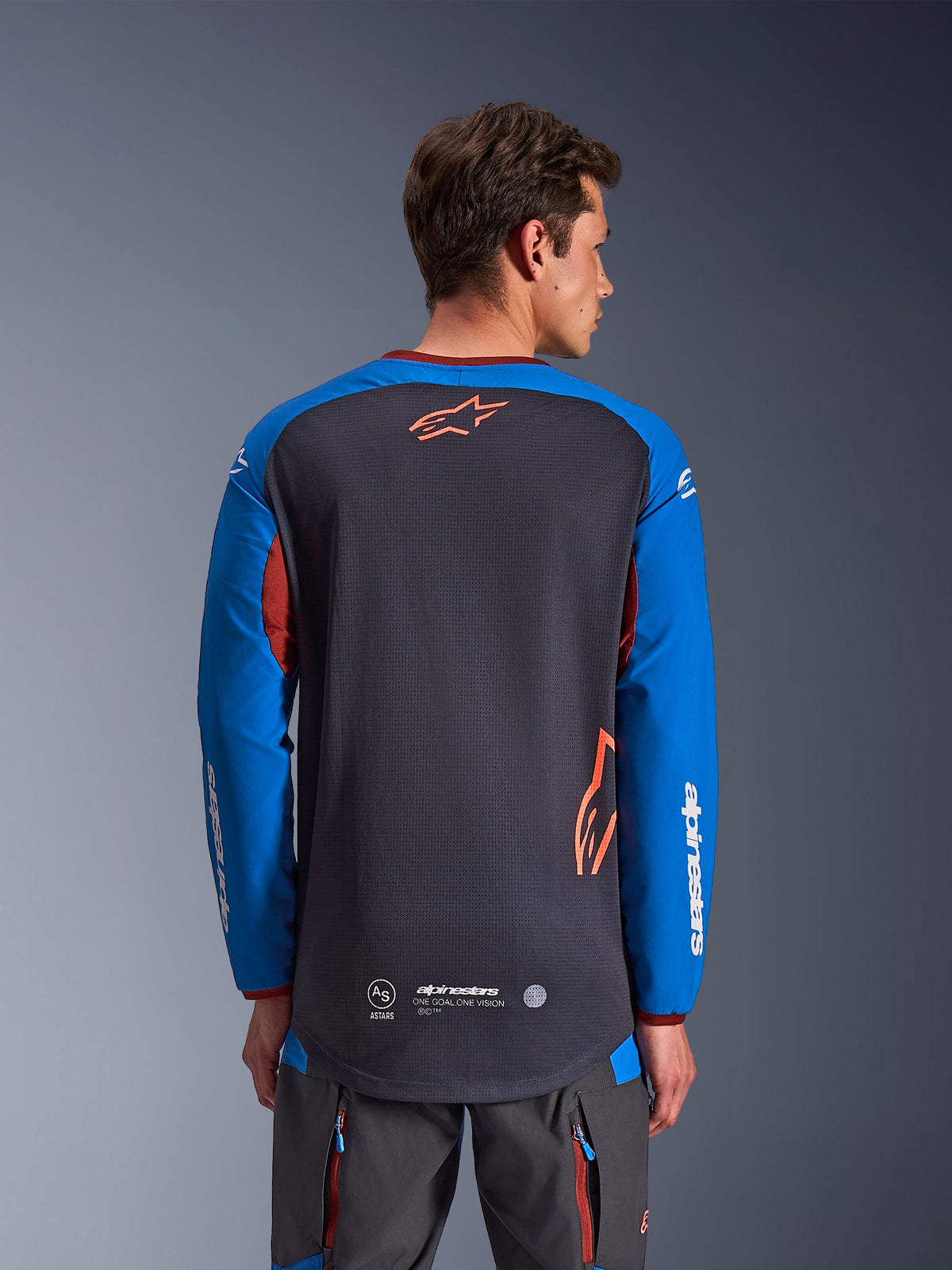 Alpinestars Maxdura Dual Jersey in Grigio Profondo e Blu Royal, vista posteriore di un modello uomo, costruzione del busto in rete con logo Astars arancione, maniche lunghe con branding bianco, colletto e polsini bordeaux, indossata con pantaloni tecnici grigi, abbigliamento da moto dual-sport e adventure.