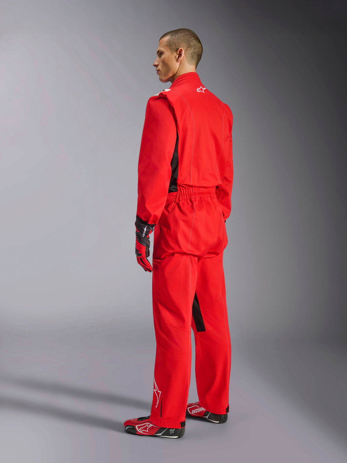 Alpinestars Kart Indoor V2 Suit, tuta da kart rossa, vista posteriore di tre quarti su un modello, indossata con guanti rossi coordinati e scarpe da corsa, costruzione in twill di policotone a strato singolo, equipaggiamento professionale da motorsport per il karting indoor.