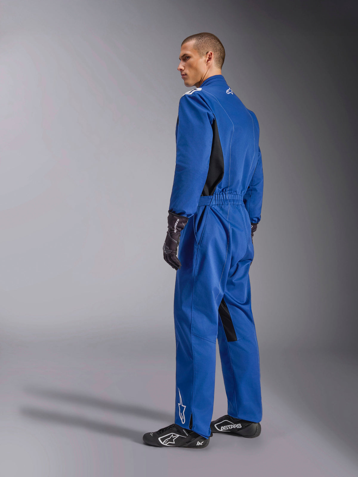Alpinestars Kart Indoor V2 Suit, tuta da kart blu, vista posteriore e laterale, tuta da corsa intera con pannelli laterali neri e inserti nell'interno gamba, vita elastica e loghi bianchi, indossata con guanti da corsa e calzature neri