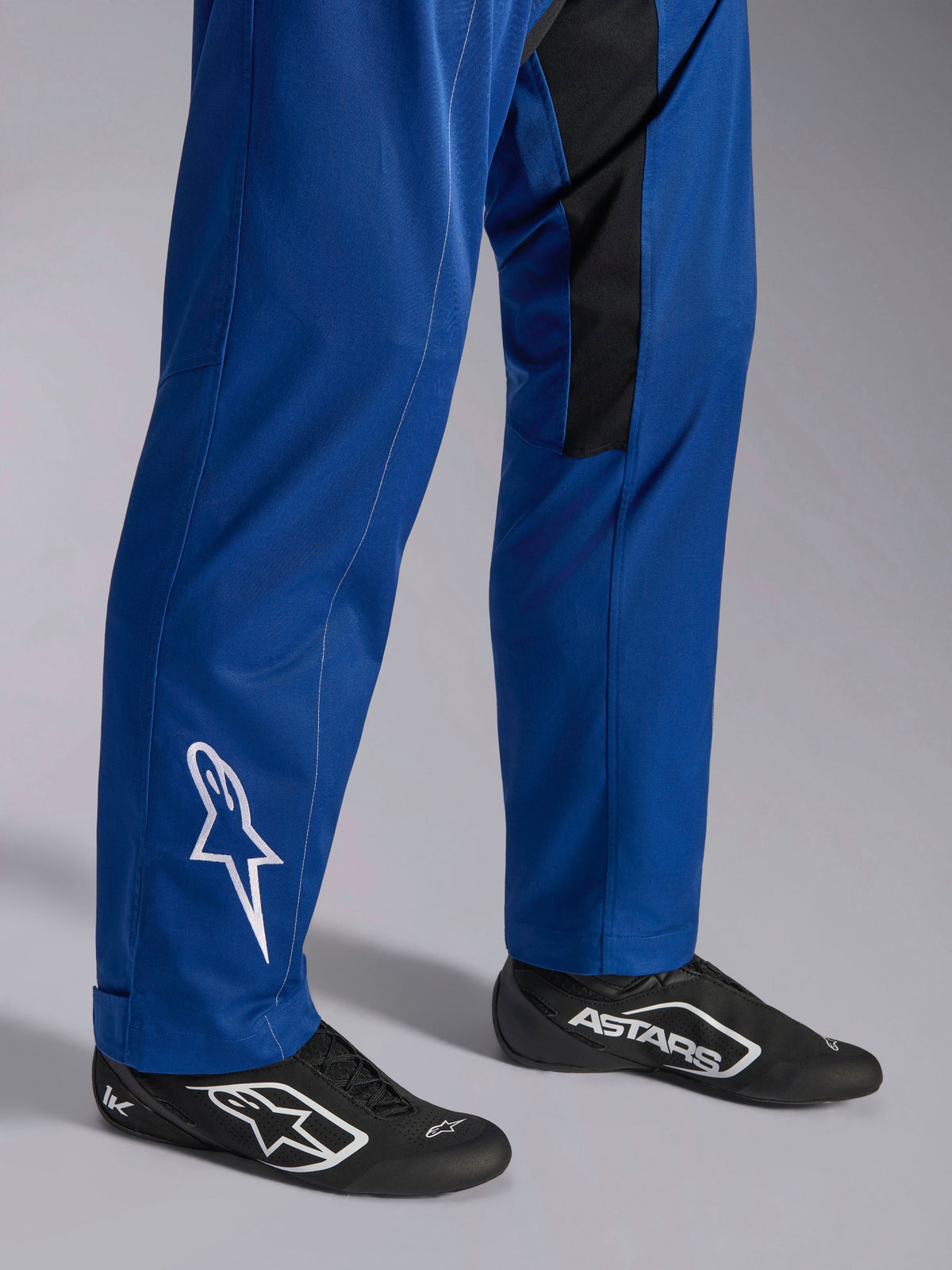 Tuta Alpinestars Kart Indoor V2, blu, primo piano delle gambe e dei piedi con logo a stella bianco sulla caviglia, fondo gamba regolabile e scarpe da corsa nere, abbigliamento da kart resistente per uso ricreativo in pista