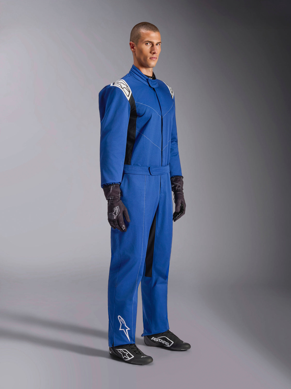 Alpinestars Kart Indoor V2 Suit, tuta da kart blu, tuta da gara intera con pannelli sulle spalle e loghi bianchi, cintura regolabile in vita, logo bianco sulla parte inferiore della gamba, indossata da un modello con guanti e scarpe da corsa neri, abbigliamento motorsport per karting indoor