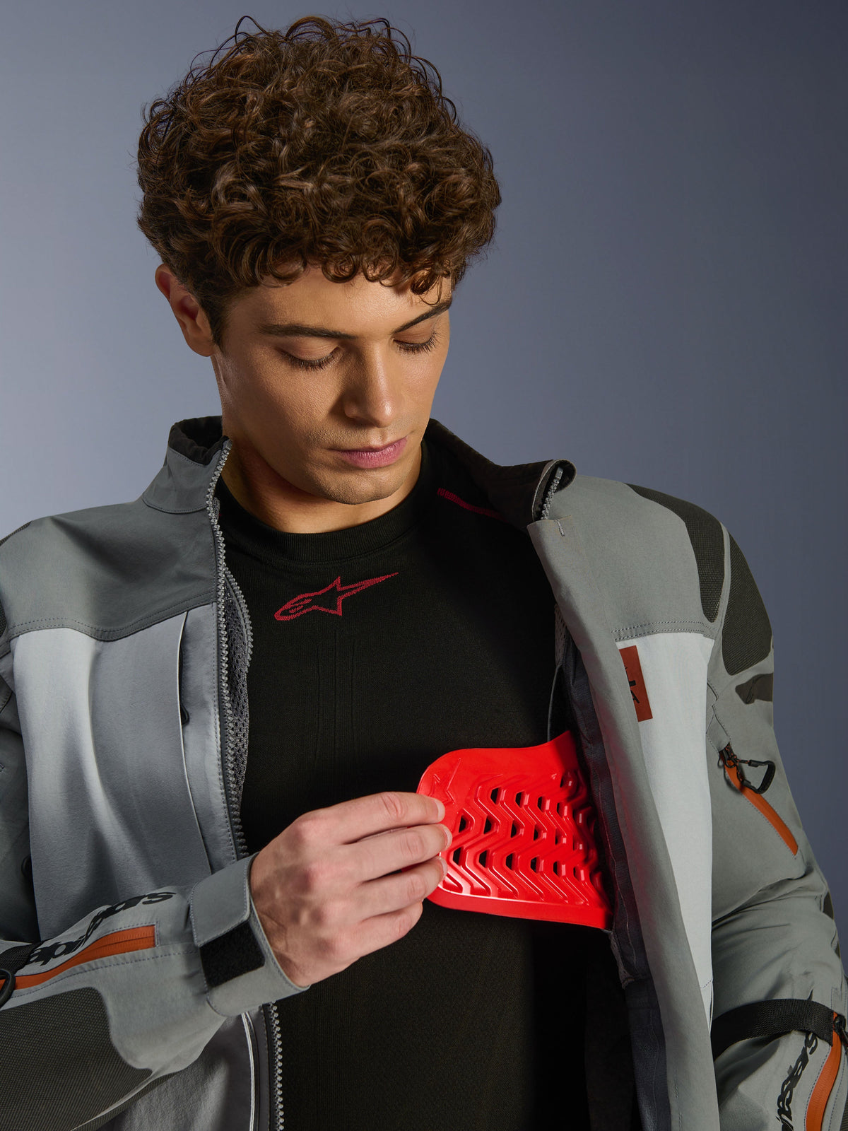 Modello che indossa la giacca Alpinestars Nazca 3L GORE-TEX PRO, grigio scuro e grigio medio, mostra l'inserto protettivo Nucleon PLASMA Pro rosso, abbigliamento moto adventure, giacca da moto premium con dettagli tecnici