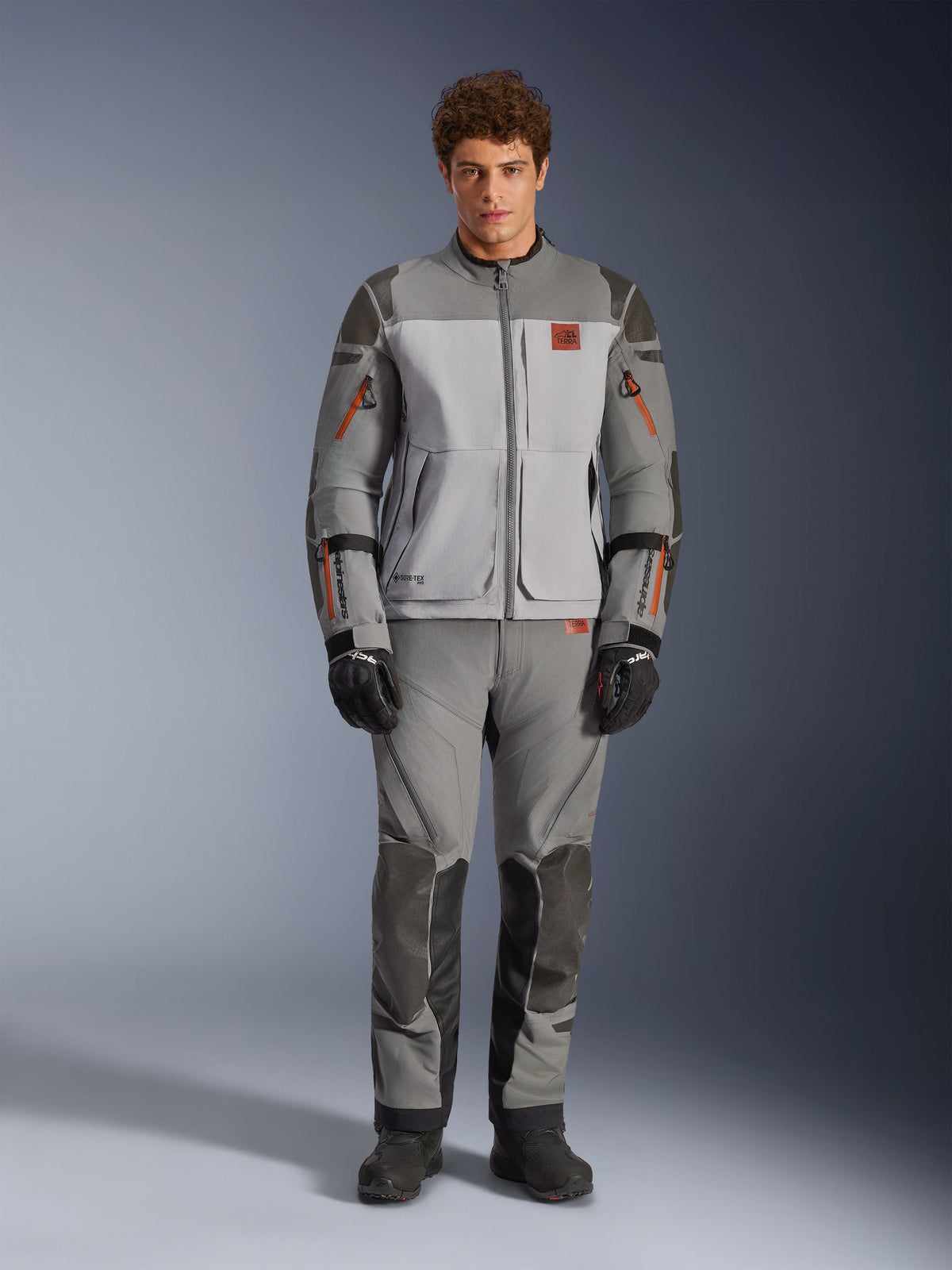 Giacca Alpinestars Nazca 3L GORE-TEX PRO, giacca da moto adventure, grigio scuro e grigio medio, indossata da un modello con pantaloni coordinati e guanti neri, con dettagli delle cerniere arancioni e logo rosso sul petto, abbigliamento tecnico protettivo per guida estrema