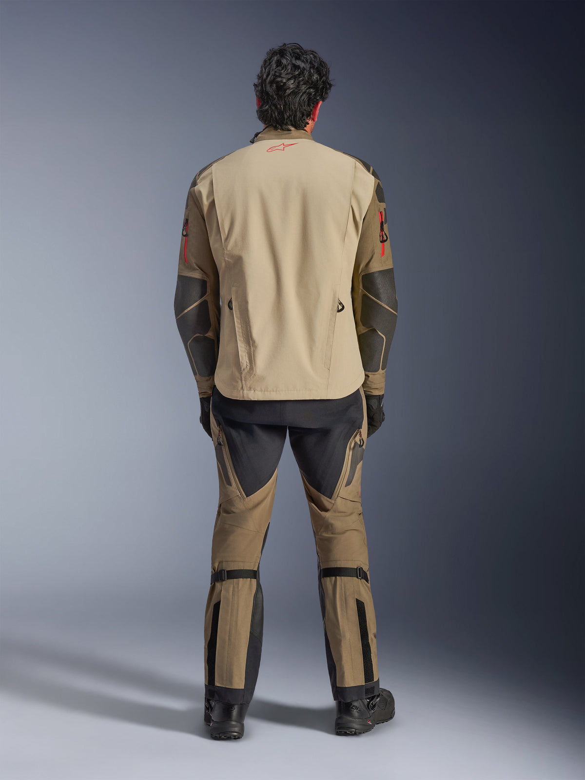 Giacca Alpinestars Nazca 3L GORE-TEX PRO in Walnut Rock Brown, vista posteriore del pilota, mostra i rinforzi in Superfabric sui gomiti e le cerniere di ventilazione posteriori, indossata con pantaloni adventure coordinati, stivali e guanti neri, abbigliamento tecnico da moto robusto per avventure estreme.