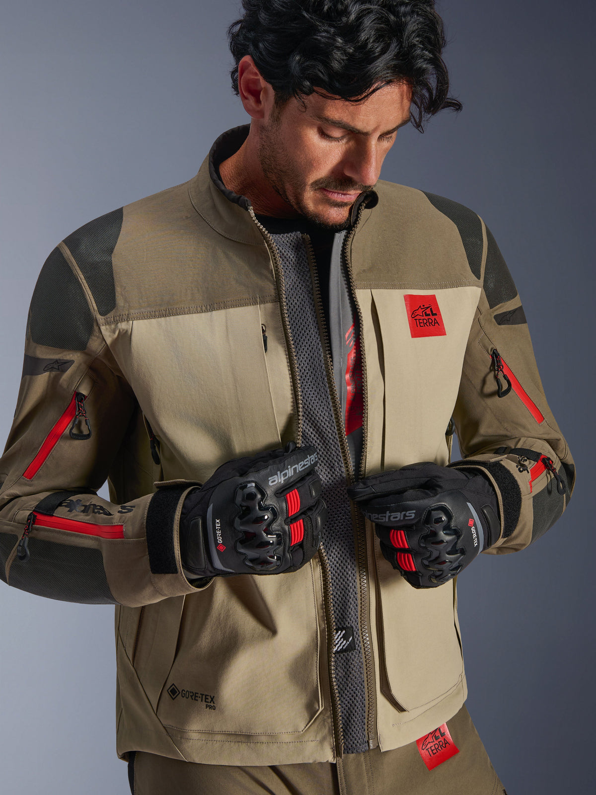 Giacca Alpinestars Nazca 3L GORE-TEX PRO, Walnut Rock Brown, giacca da moto adventure con parte anteriore aperta che rivela il pannello centrale in mesh per il flusso d'aria, logo All Terra rosso, indossata con guanti da moto GORE-TEX neri, equipaggiamento tecnico per touring fuoristrada
