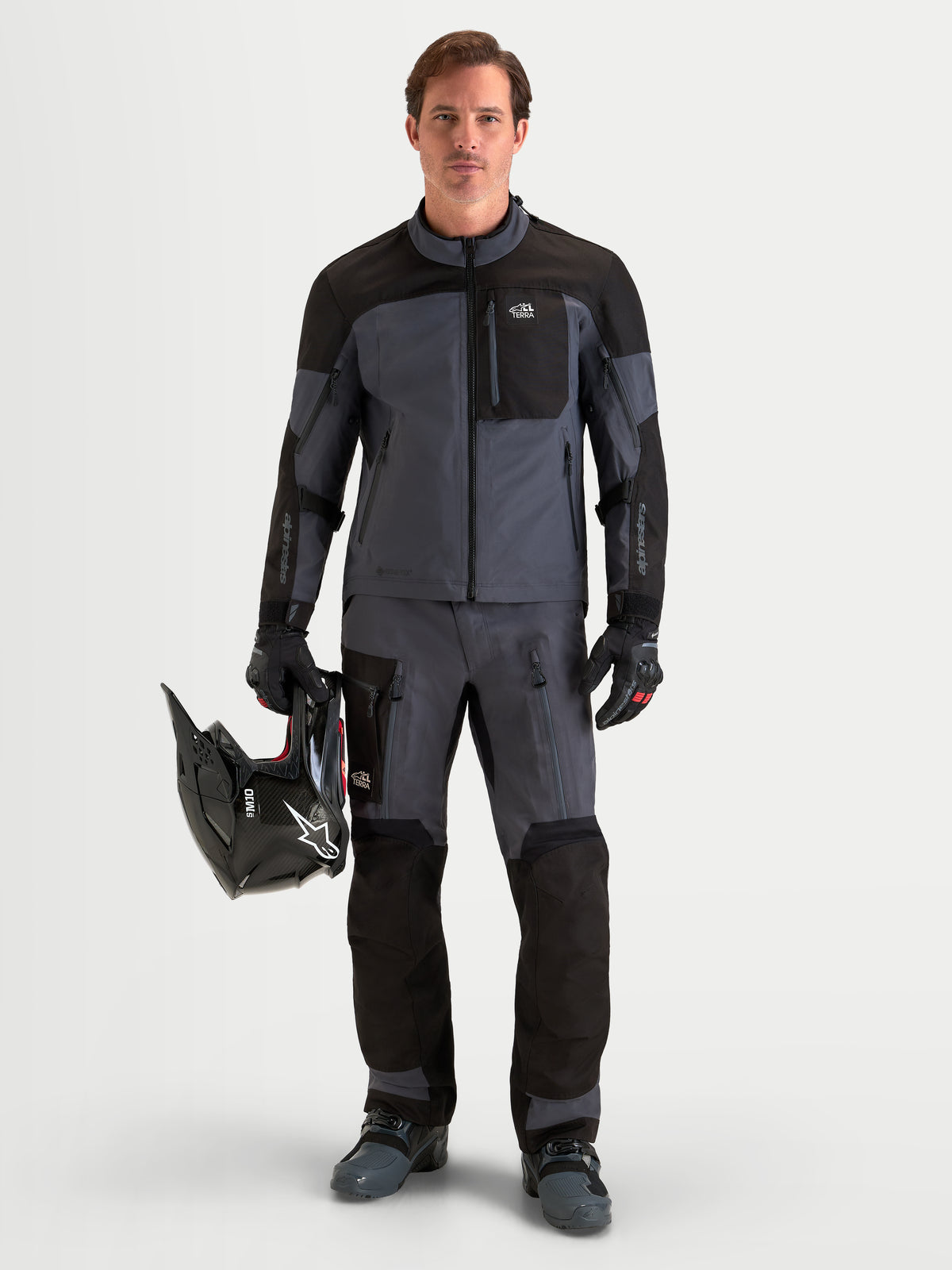 Alpinestars Arenis Gore-Tex Gear Set, completo da moto adventure da uomo in nero e grigio scuro, vista frontale di un modello in piedi che indossa abbigliamento tecnico impermeabile coordinato e tiene in mano un casco