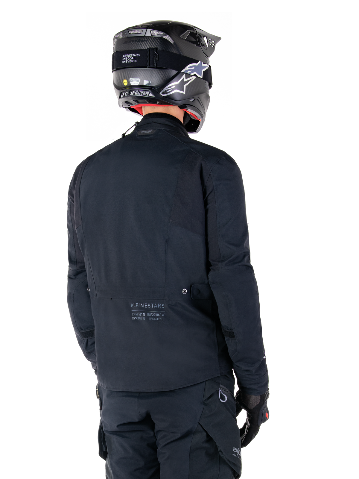Giacca Alpinestars St-7 Prime GORE-TEX, nero, vista posteriore di un pilota che indossa la giacca con casco e pantaloni coordinati, con marchio Alpinestars e coordinate geografiche sulla parte bassa della schiena.