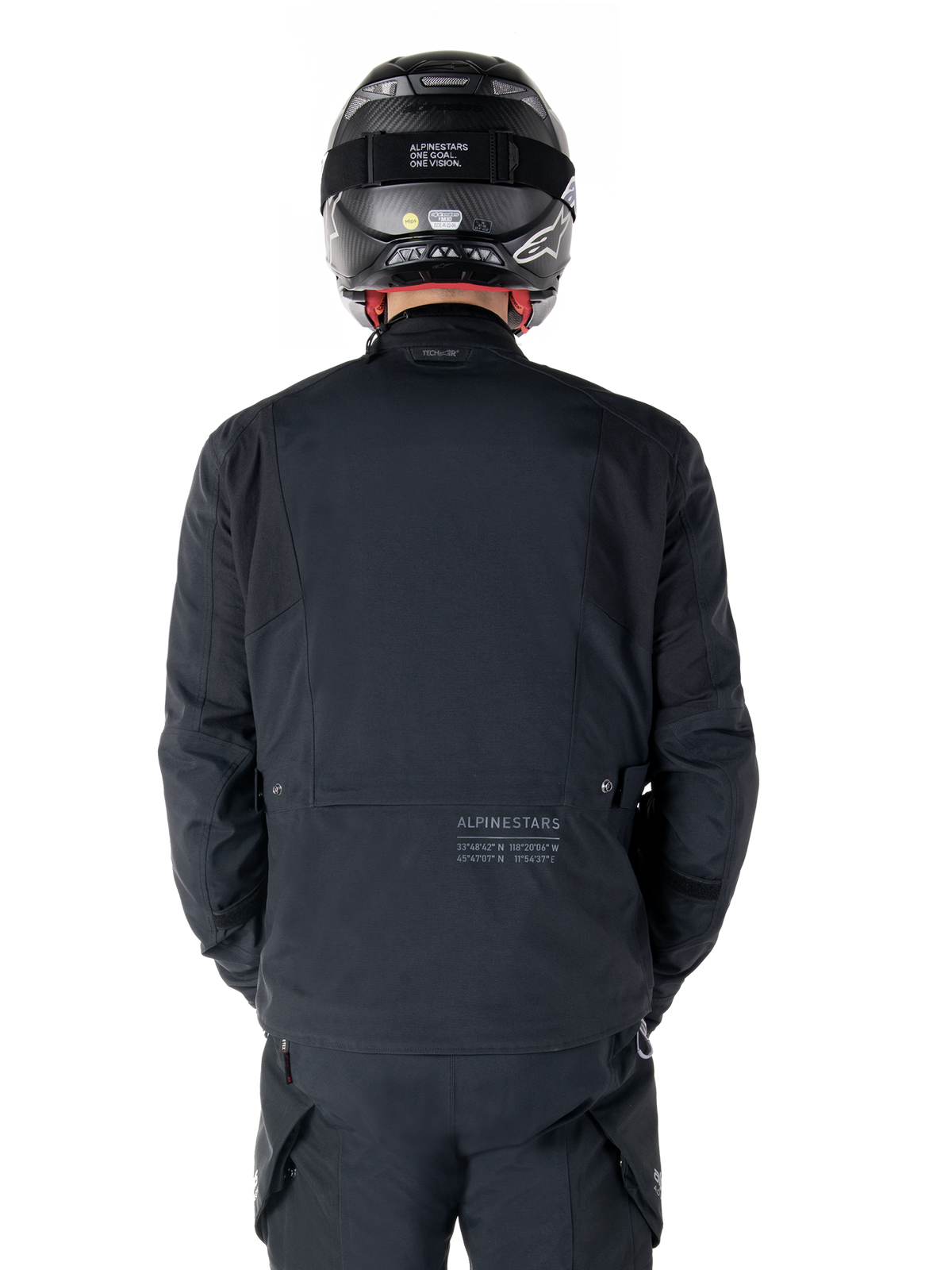 Giacca Alpinestars St-7 Prime GORE-TEX, nera, vista posteriore di un motociclista, con logo discreto e coordinate geografiche sulla parte bassa della schiena, indossata con casco e pantaloni neri coordinati, abbigliamento tecnico da motociclismo