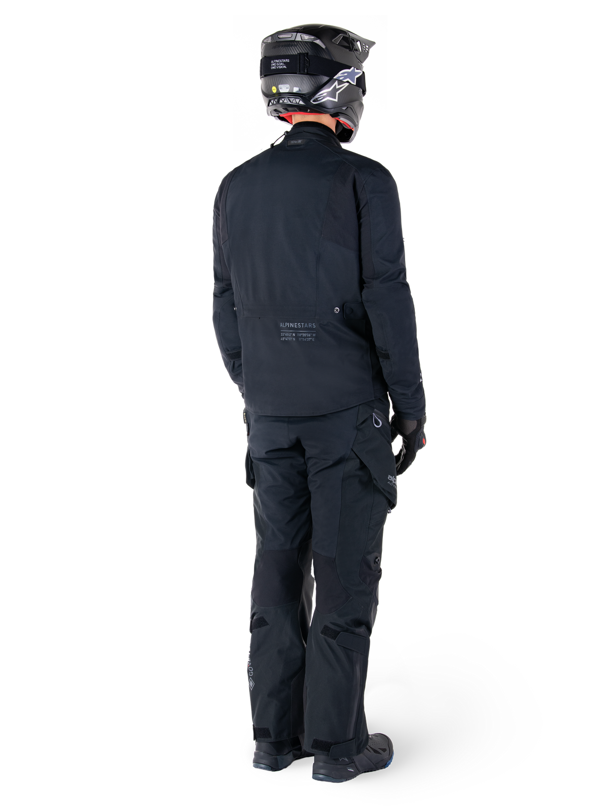 Vista posteriore di un pilota con giacca Alpinestars St-7 Prime GORE-TEX nera, pantaloni da touring neri coordinati, casco da moto nero con logo bianco e stivali neri, abbigliamento protettivo adventure completo per touring e commuting.