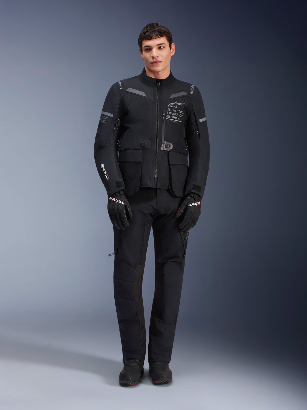 Giacca Alpinestars ST-7 2L GORE-TEX, giacca da moto adventure touring, nero e grigio scuro, indossata con pantaloni e guanti da moto neri coordinati, con tasche cargo multiple, inserti riflettenti e logo Alpinestars Exploration Equipment sul petto, design funzionale per la guida su strada e fuoristrada
