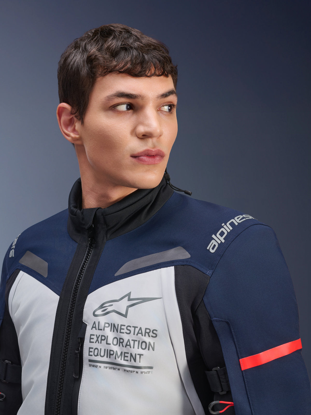 Giacca Alpinestars ST-7 2L GORE-TEX, grigio ghiaccio, blu scuro e nero, indossata da un modello, con logo Alpinestars Exploration Equipment sul petto, collo alto e dettaglio rosso sulla manica, design in tessuto resistente per adventure touring