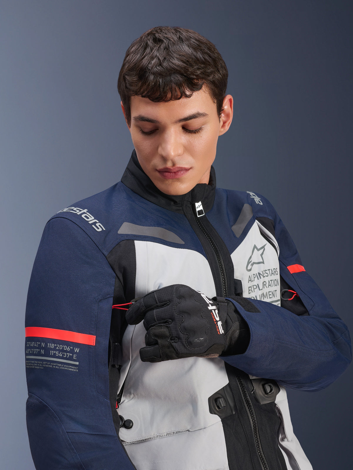 Giacca Alpinestars ST-7 2L GORE-TEX, Ice Gray, Blu Scuro e Nero con accenti arancioni, indossata da un uomo che regola un dettaglio della manica, con marchio 'Alpinestars Exploration Equipment' e coordinate geografiche, abbigliamento professionale da moto adventure
