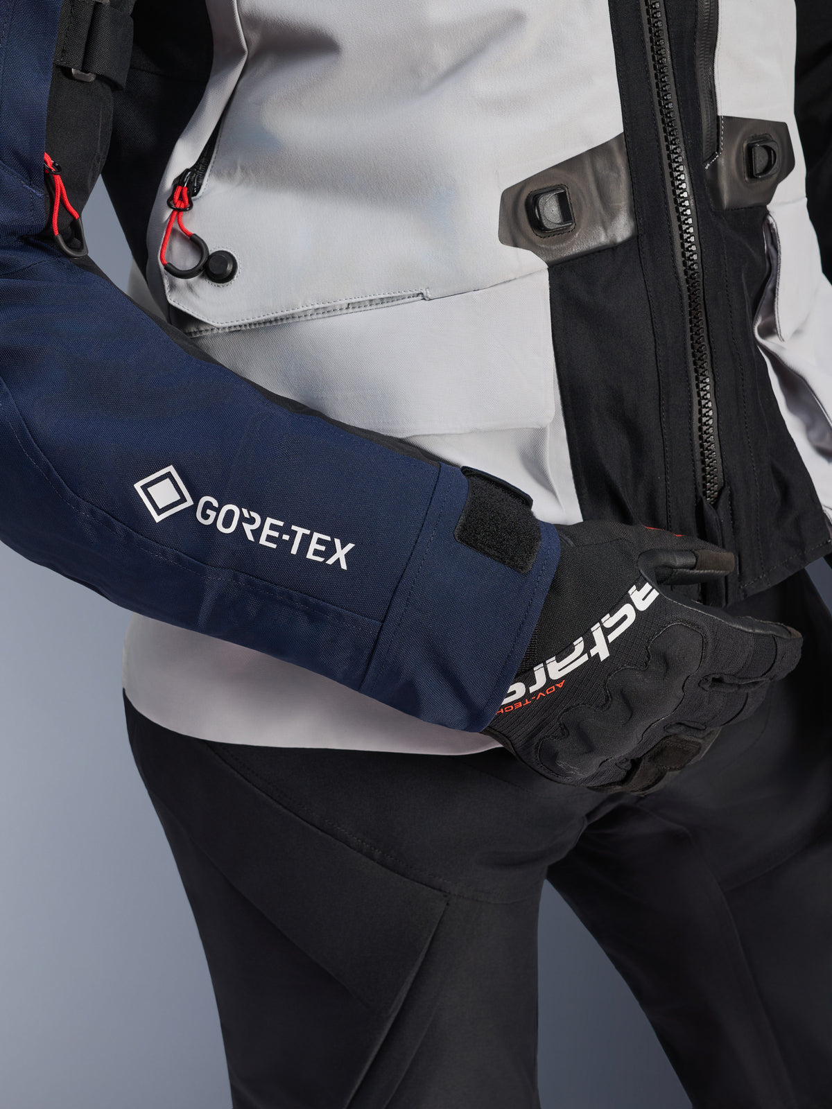 Giacca Alpinestars ST-7 2L GORE-TEX, primo piano della manica blu scuro con logo GORE-TEX bianco, busto grigio ghiaccio con dettagli neri e accenti in cordino rosso, indossata con guanti e pantaloni neri Alpinestars, abbigliamento da moto adventure touring