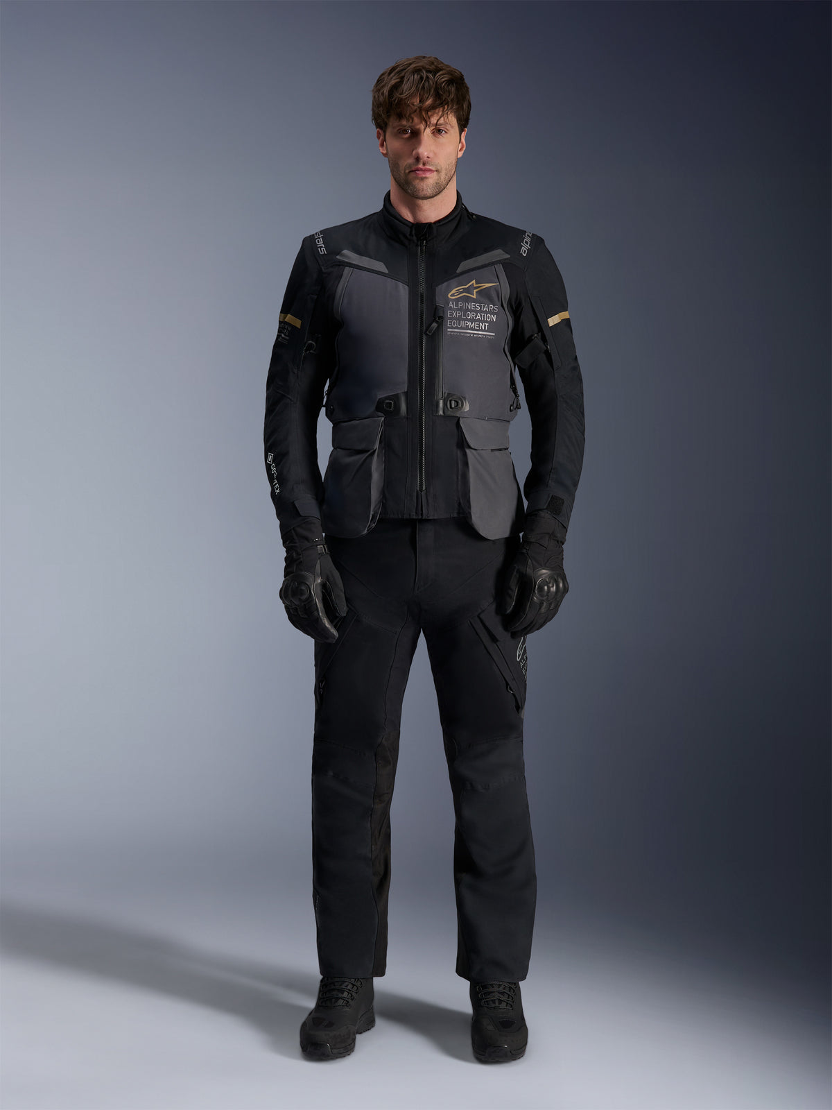Modello che indossa la giacca Alpinestars ST-7 2L GORE-TEX in Deep Gray Black Dark Khaki, con il marchio 'ALPINESTARS EXPLORATION EQUIPMENT' sul petto, abbinata a pantaloni da moto, guanti e stivali neri coordinati, vista frontale in studio.
