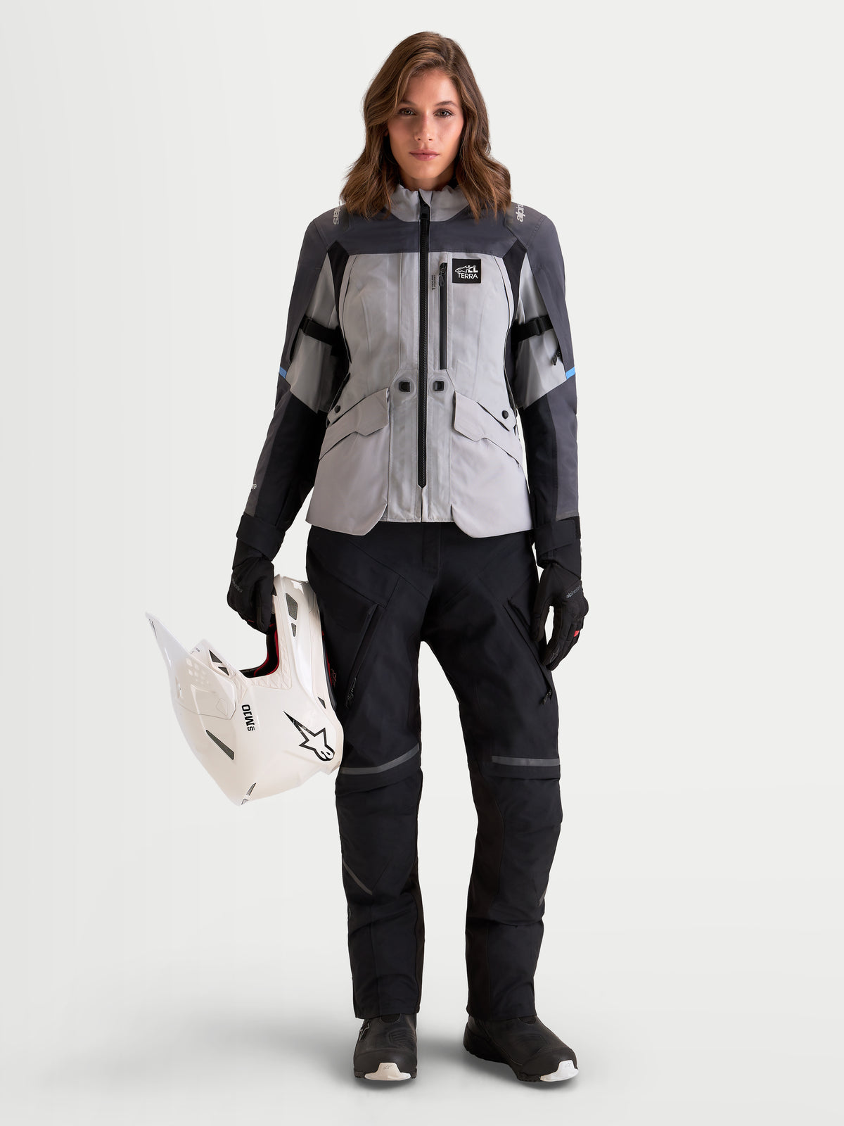 Alpinestars Stella ST-7 V2 2L Gore-Tex Giacca, giacca da moto adventure da donna in grigio medio, grigio scuro e nero, vista frontale indossata da una modella che tiene un casco da off-road bianco, con struttura in tessuto impermeabile e certificazione CE