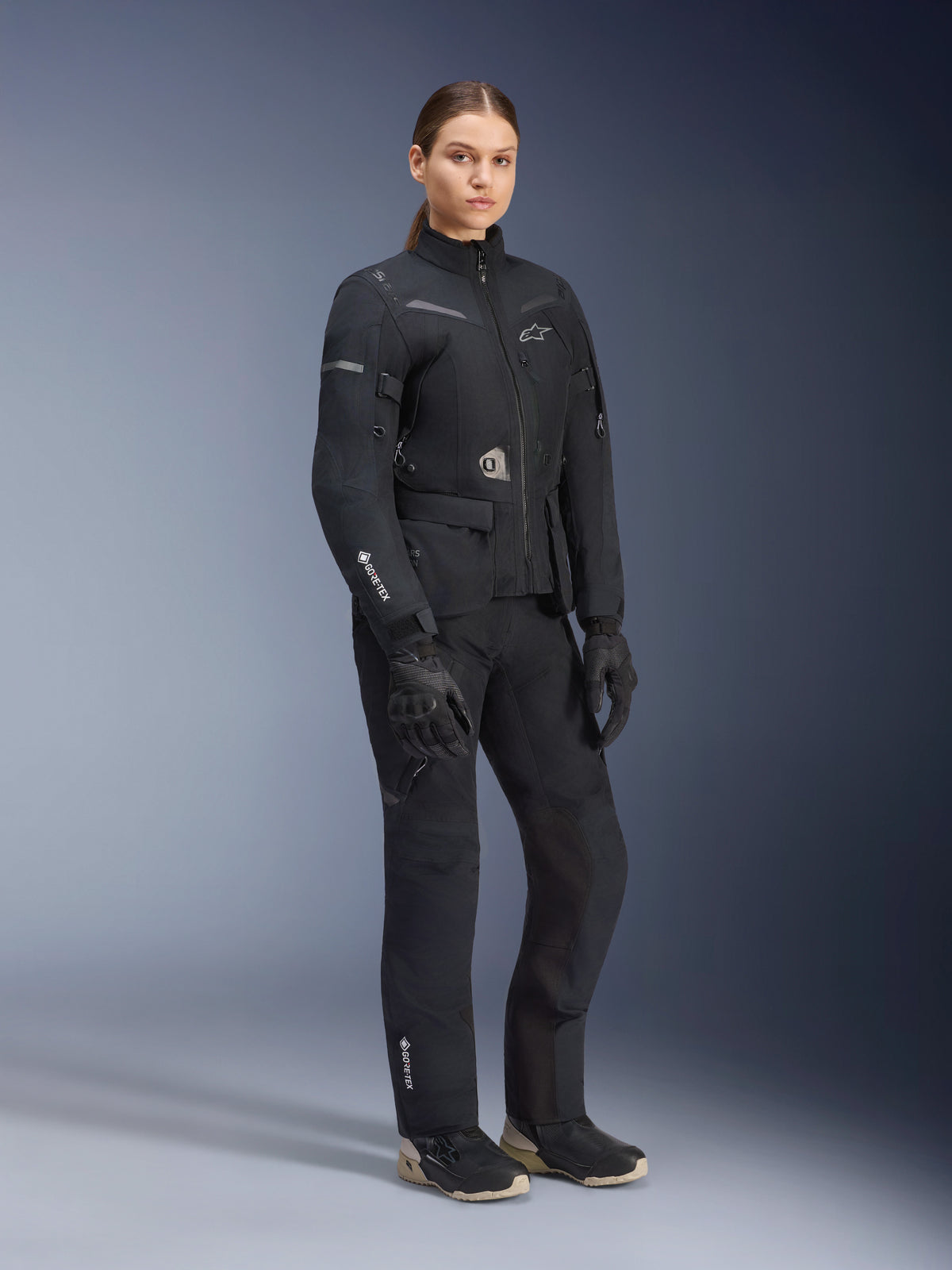Modella che indossa la tuta adventure touring Alpinestars Stella ST-7 2L GORE-TEX, giacca e pantaloni nero e grigio scuro, con marchio GORE-TEX in evidenza su manica e gamba, abbinata a guanti neri e stivali da moto, equipaggiamento professionale da motociclismo per ogni condizione meteo
