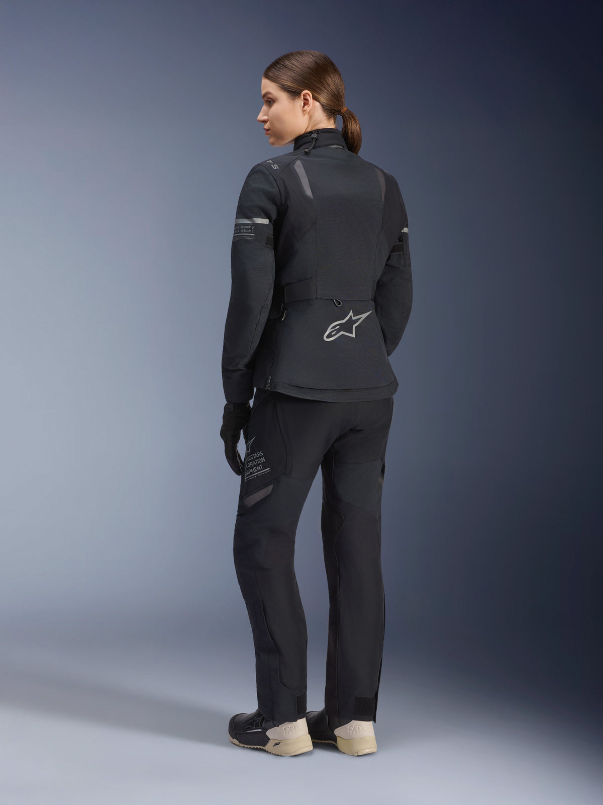 Giacca e pantaloni da donna Alpinestars Stella ST-7 2L GORE-TEX, completo da touring adventure completamente nero, vista posteriore che mostra il logo a stella Alpinestars e il marchio Exploration Equipment, abbinati a stivali beige e guanti neri.