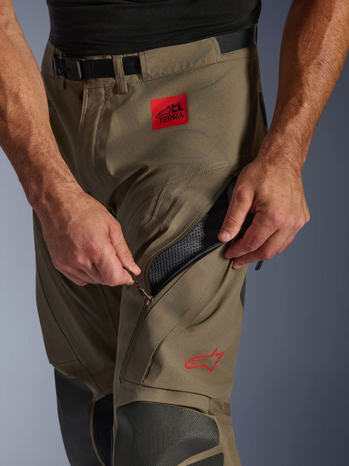 Pantaloni Alpinestars Nazca 3L GORE-TEX PRO in Walnut Black, dettaglio ravvicinato della zona della coscia che mostra una mano che apre la grande presa d'aria con cerniera, con patch rossa ALL TERRA e logo Alpinestars rosso, abbigliamento tecnico da moto adventure.