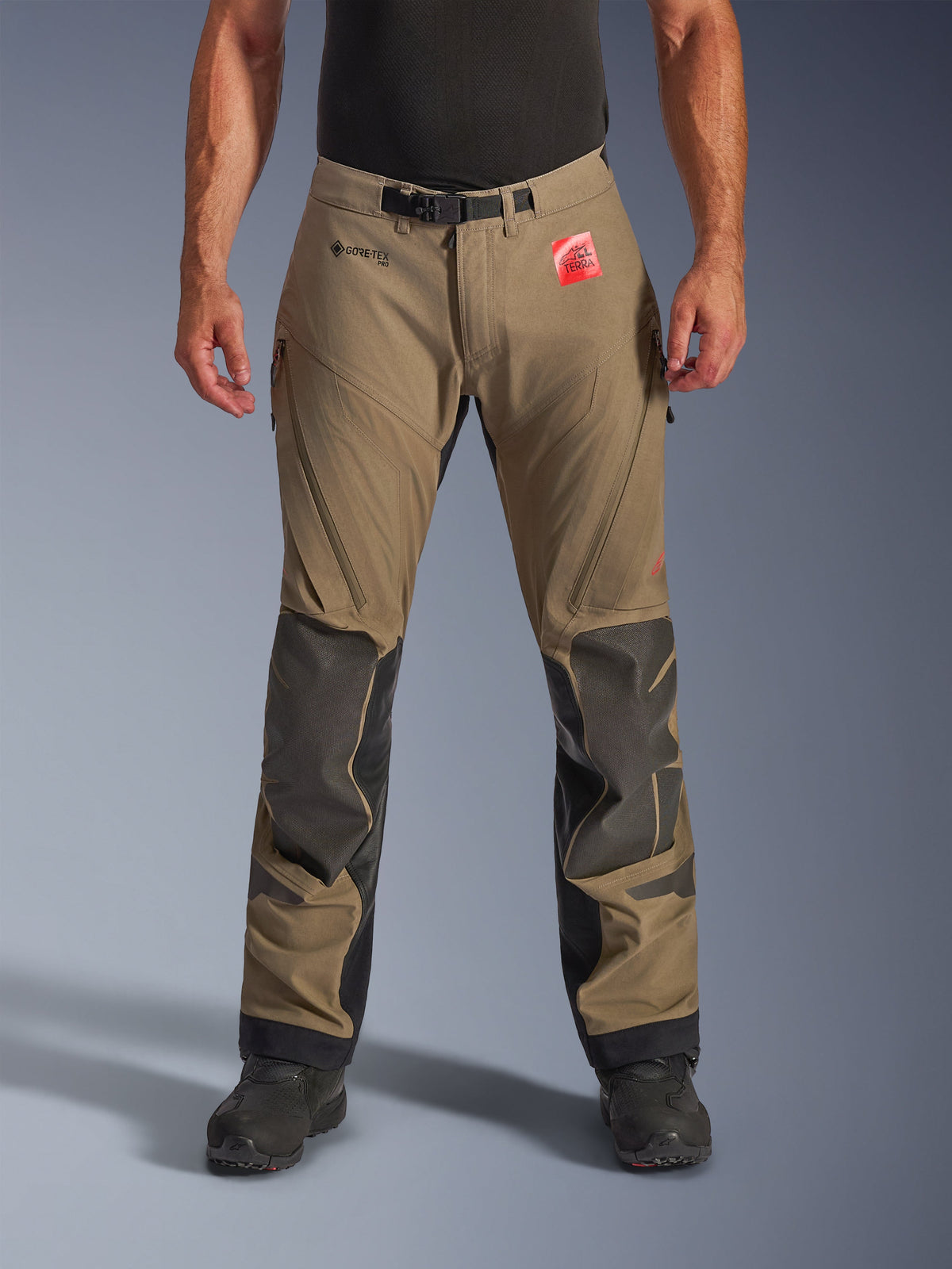Alpinestars Nazca 3L GORE-TEX PRO Pants in Walnut Black, pantaloni da moto adventure touring da uomo, con tessuto marrone e rinforzi sulle ginocchia in Superfabric nero, prese d'aria con zip sulle cosce e chiusura magnetica in vita, vista frontale su modello con stivali neri, abbigliamento tecnico impermeabile