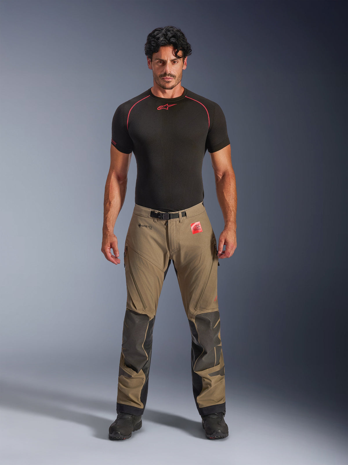 Modello che indossa pantaloni Alpinestars Nazca 3L GORE-TEX PRO in marrone noce e nero, abbigliamento tecnico da moto adventure con rinforzi sulle ginocchia in Superfabric e fibbia magnetica in vita, abbinati a una maglia base layer Alpinestars nera, vista frontale in studio.