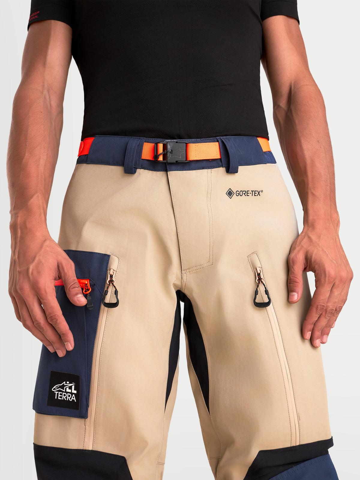 Alpinestars Arenis Gore-Tex Pantaloni, pantaloni da moto adventure da uomo in beige e blu scuro con cintura in vita arancione, primo piano che mostra il marchio GORE-TEX, fibbia magnetica, prese d'aria sulle cosce e tasca cargo indossati da un modello