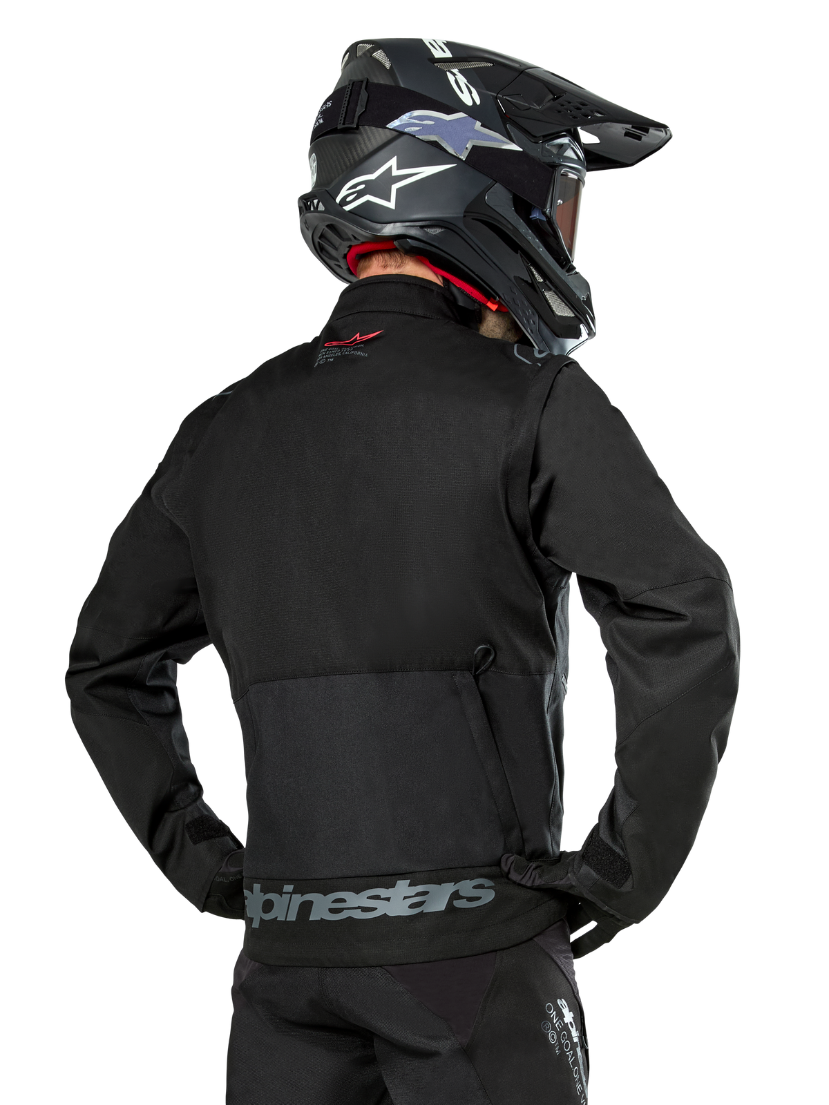 Vista posteriore di un pilota che indossa la giacca Alpinestars Pro-Dura in nero, giacca da enduro in tessuto ripstop resistente, caratterizzata da una grande tasca cargo posteriore con marchio Alpinestars grigio sulla parte bassa della schiena, abbinata a un casco da motocross nero.