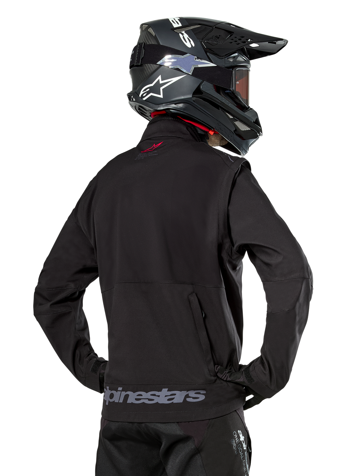 Giacca Alpinestars Lite-Dura Softshell, nero, vista posteriore indossata da un pilota, con branding Alpinestars grigio sull'orlo posteriore e un piccolo logo rosso sul colletto, abbinata a un casco da motocross nero, progettata per l'enduro e il fuoristrada