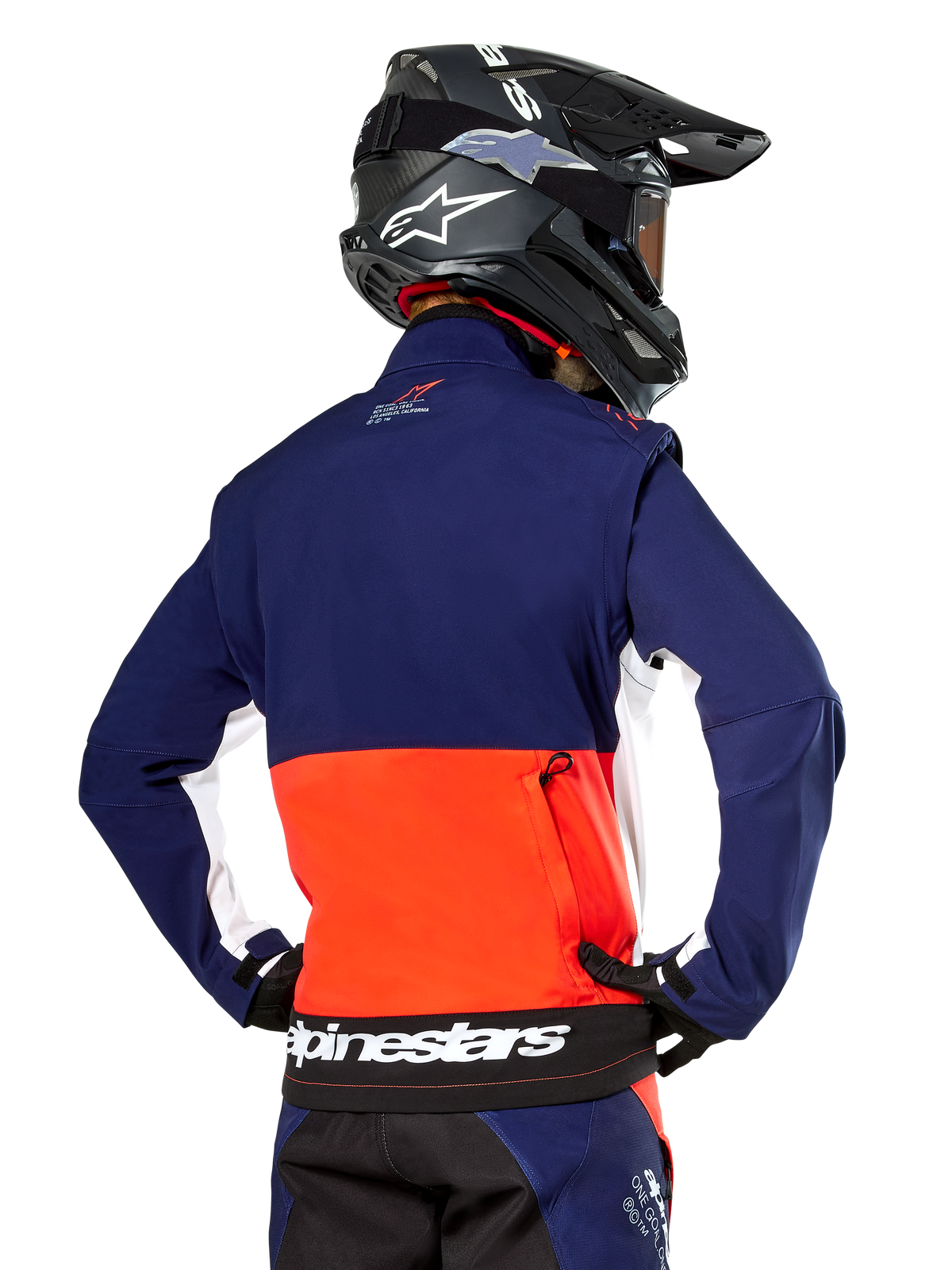 Giacca Softshell Alpinestars Lite-Dura, blu navy, arancione acceso e bianco, vista posteriore indossata da un pilota di motocross, con un grande logo bianco sull'orlo posteriore e tasca portaoggetti con zip, parte di un completo da gara off-road