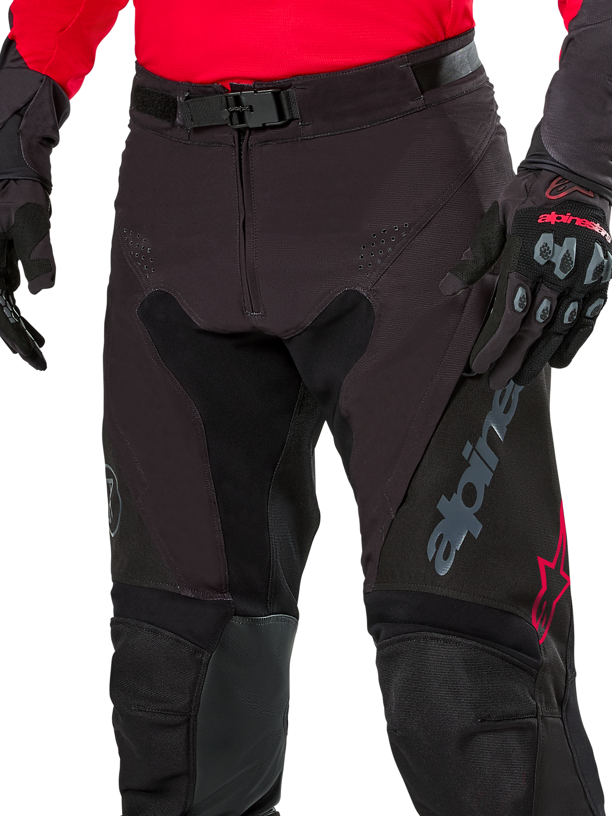 Primo piano dei pantaloni Alpinestars Pro-Dura, pantaloni da enduro, nero con accenti rossi, dotati di chiusura sicura con fibbia magnetica e patta con cerniera, indossati con maglia rossa coordinata e guanti neri, robusta costruzione in ripstop per guida off-road